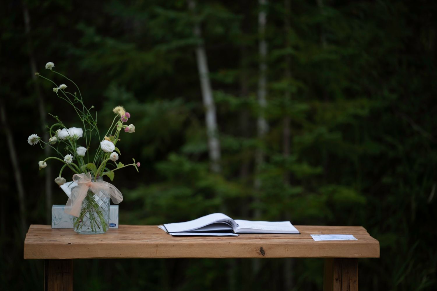 Rustic wooden table with white flowers in vases set against a dark green forest backdrop.