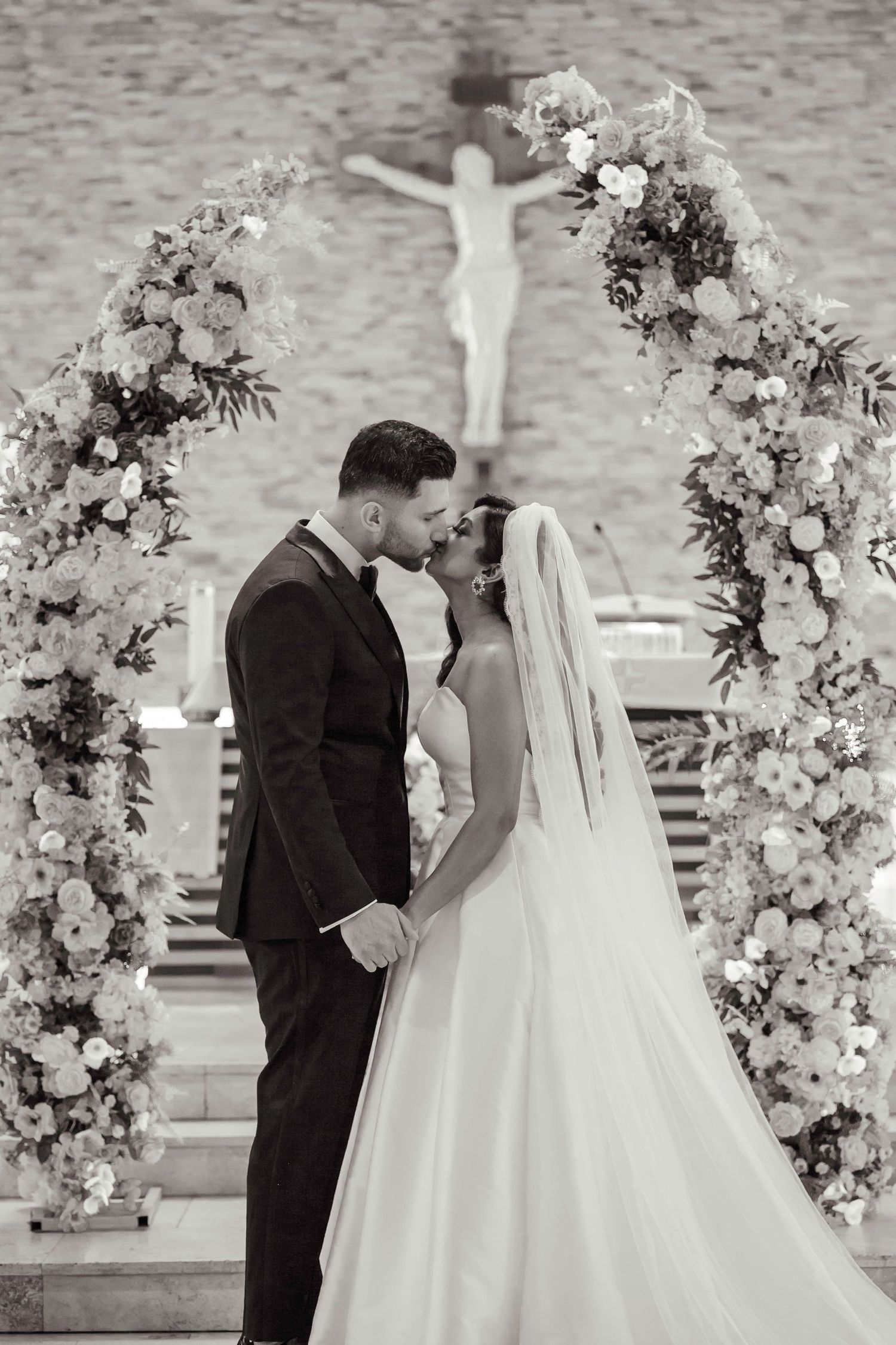 Black and white photo of couple during wedding ceremony under white floral arch.