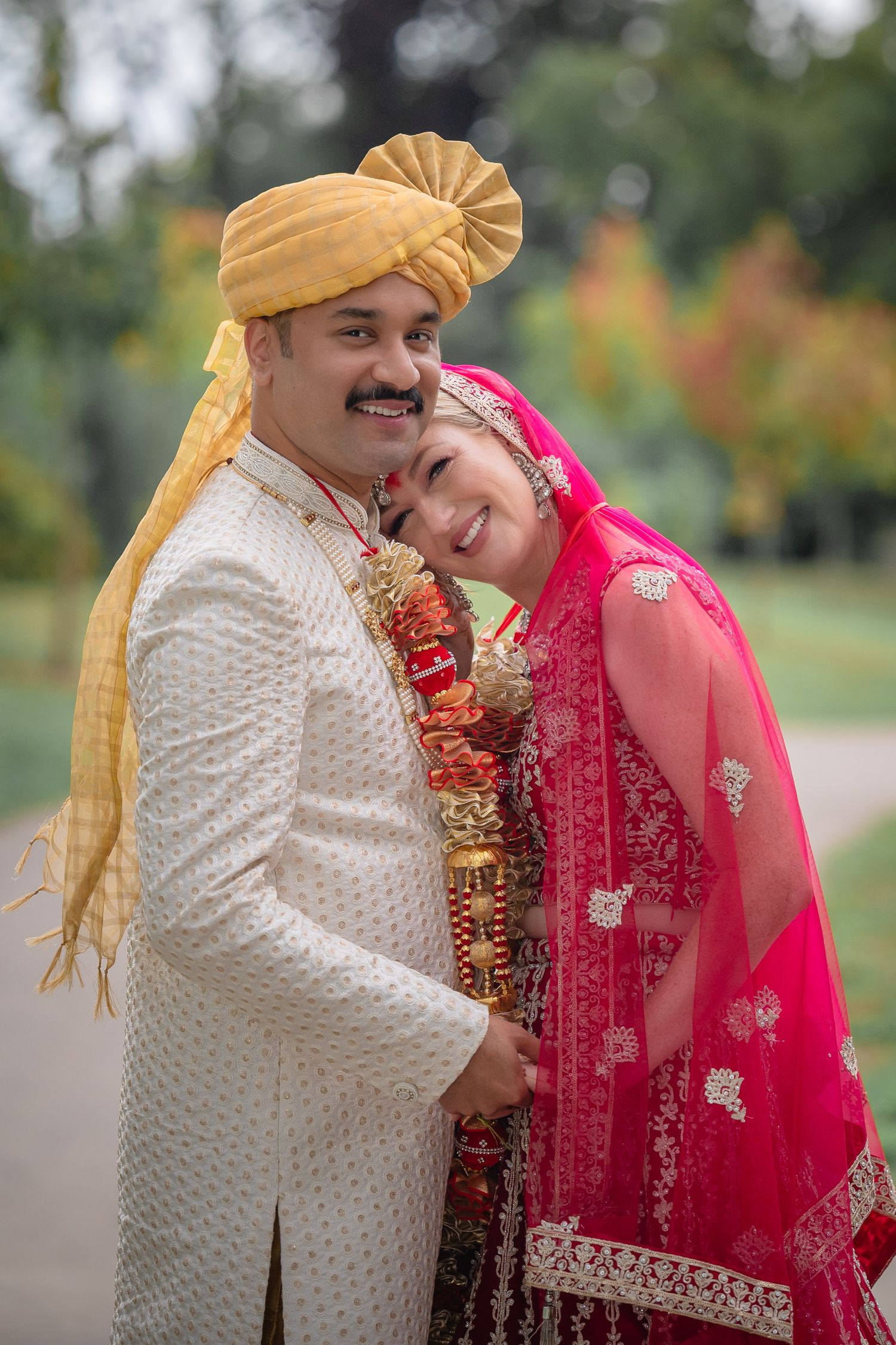 A couple in traditional Indian wedding attire share an intimate moment outdoors in their white and red outfits.