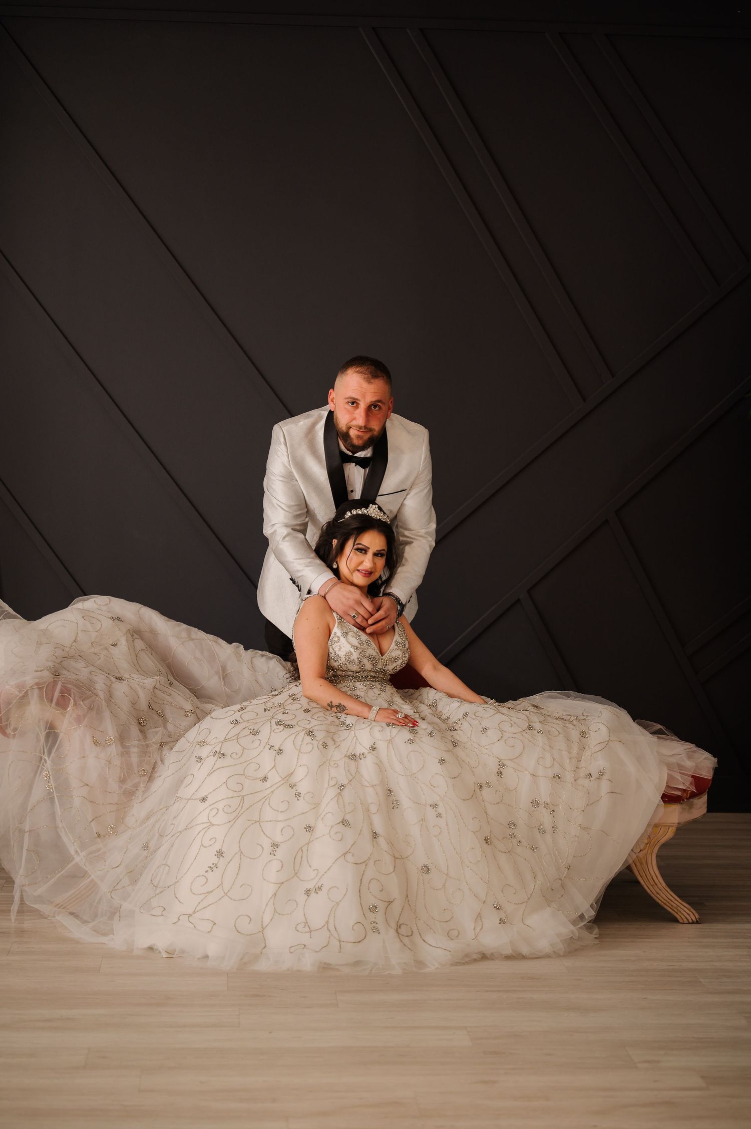 Elegant wedding couple posing on floor in formal attire against dark backdrop.