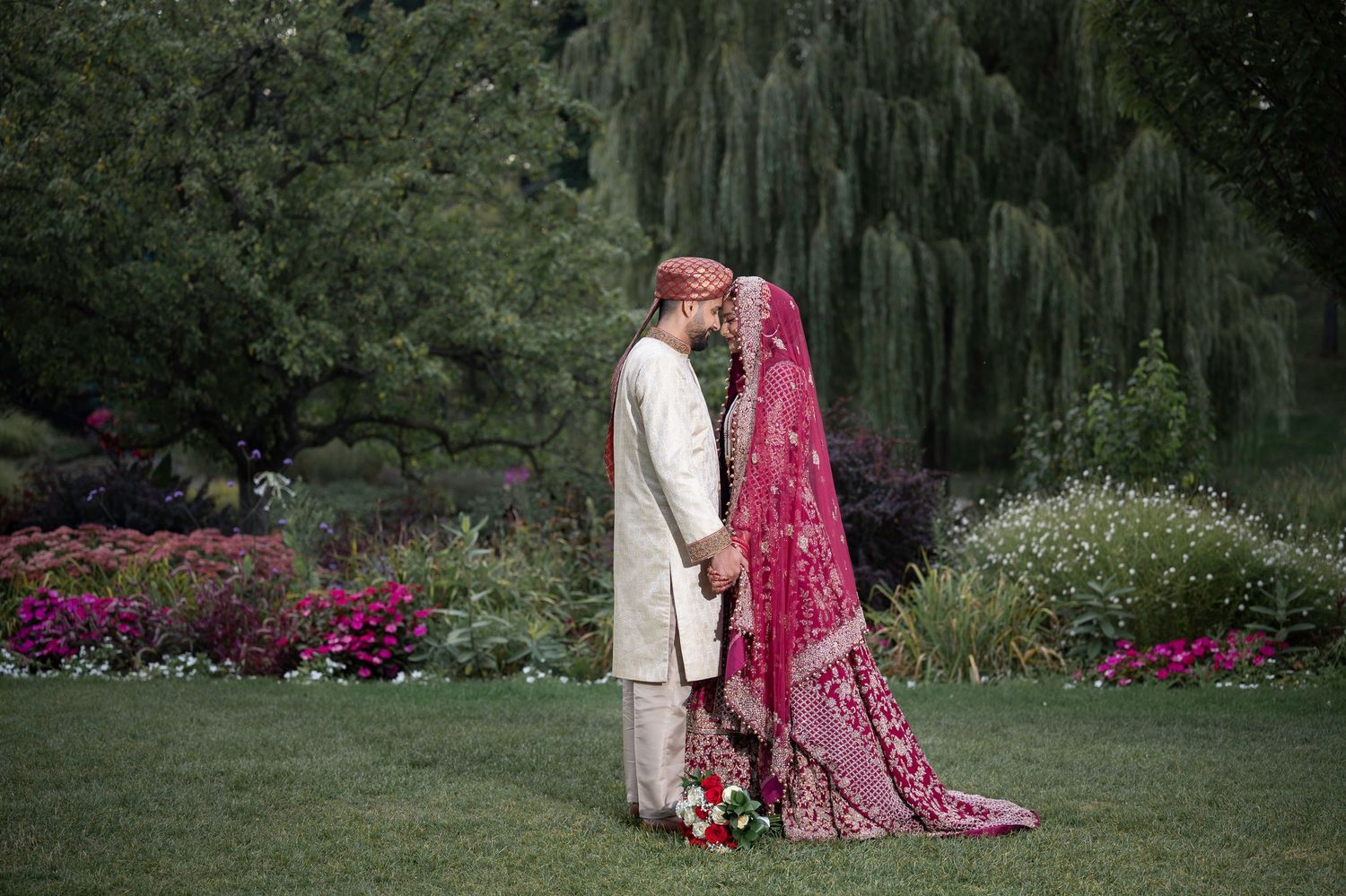 A couple poses in a garden setting with traditional wedding and cultural attire.