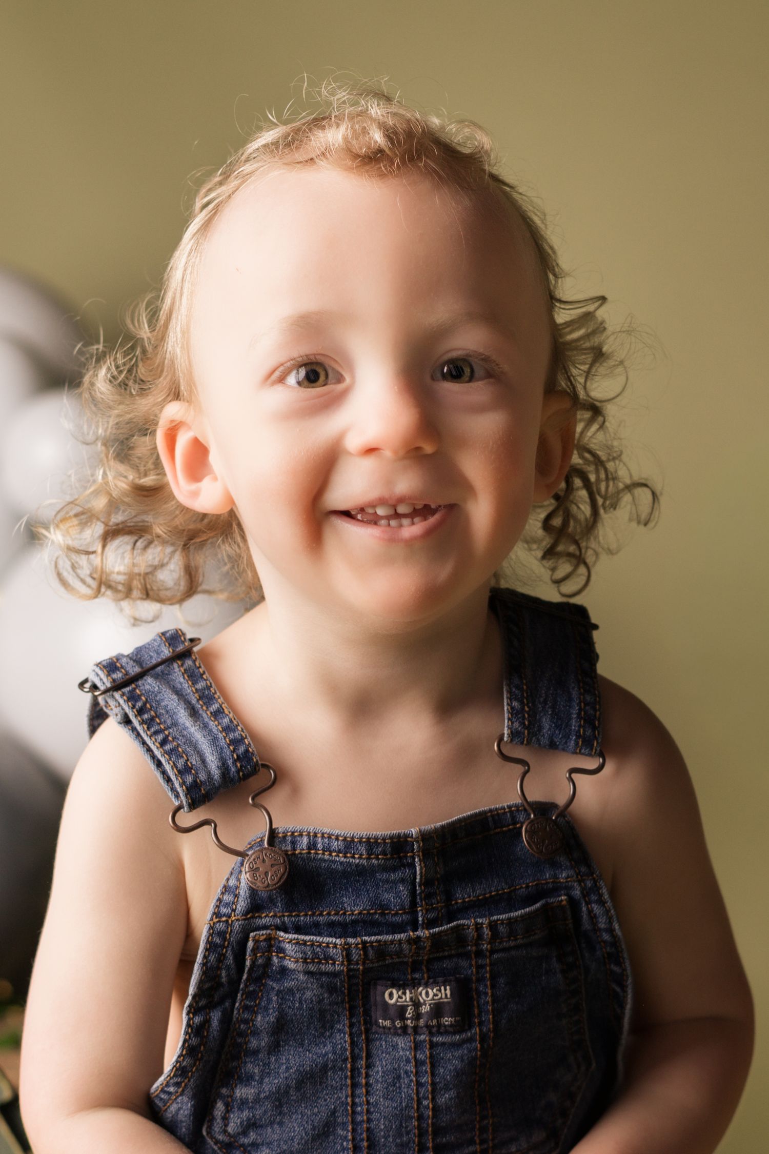 A young child wearing denim overalls smiles brightly against a warm-toned background.