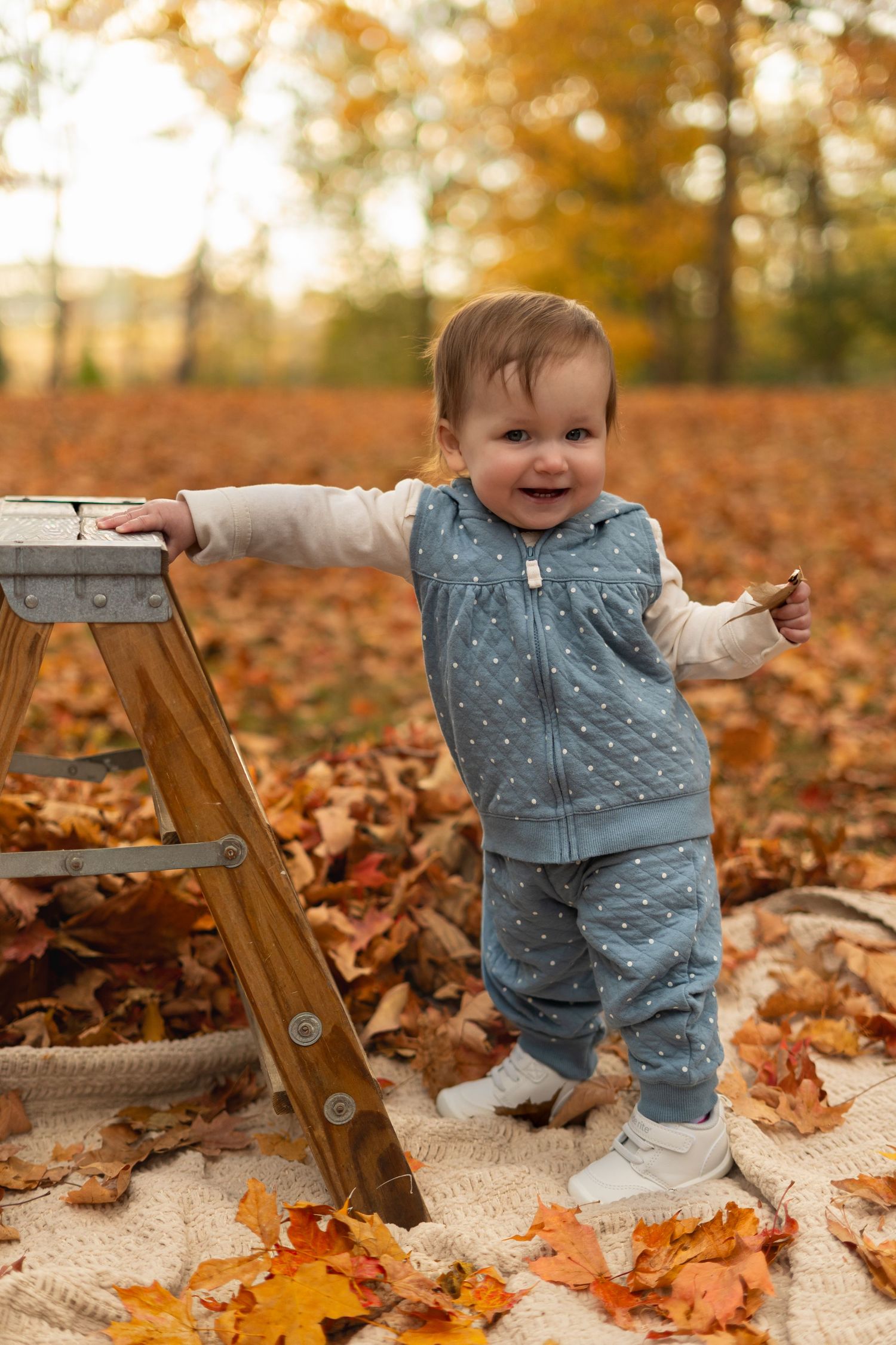 Small child in blue outfit playing among autumn leaves on a blanket outdoors.