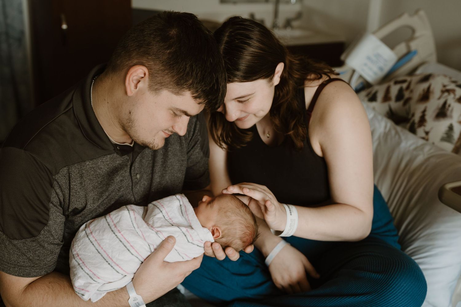 New parents sitting on hospital bed lovingly gazing at their newborn baby wrapped in white blanket during Fresh 48 in-hospital newborn photo