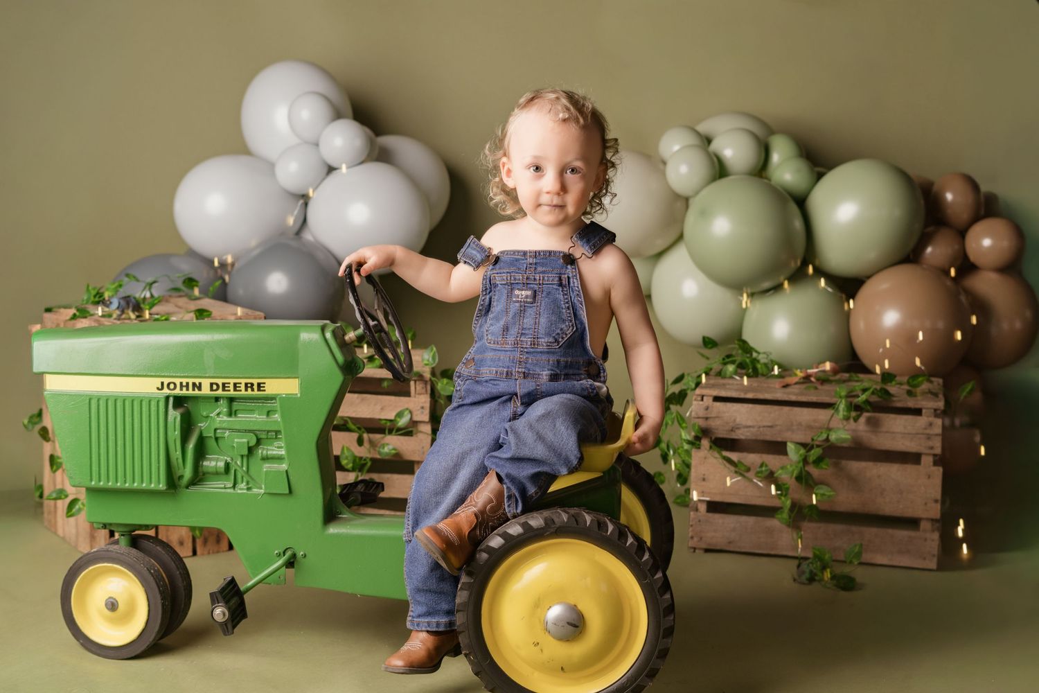 A child-sized green tractor setup with balloons and wooden crates creates a rustic photography scene.