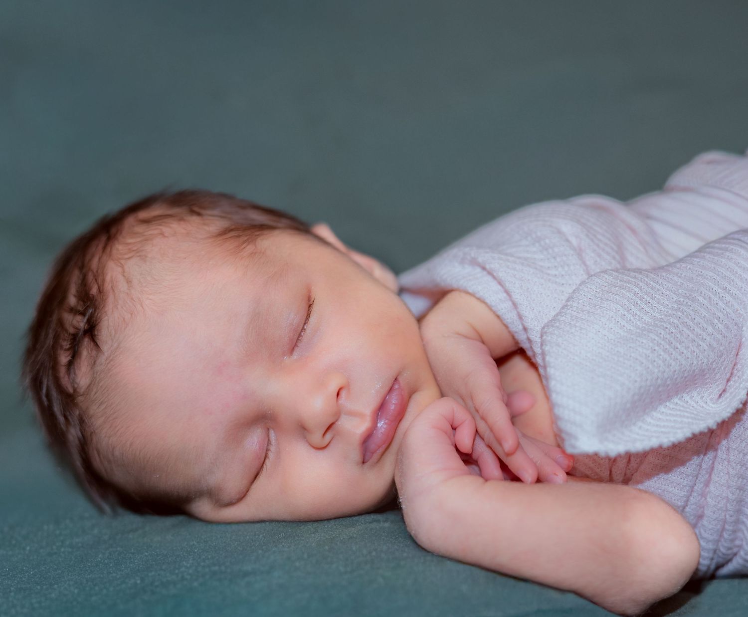 Peaceful sleeping newborn baby wearing white sweater rests on teal blanket with hands tucked under chin.