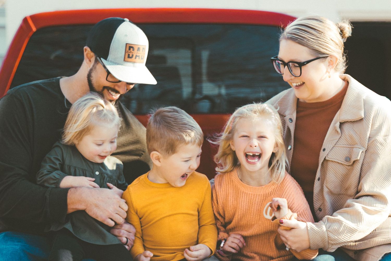 Family sharing laughs while sitting together on red truck tailgate.
