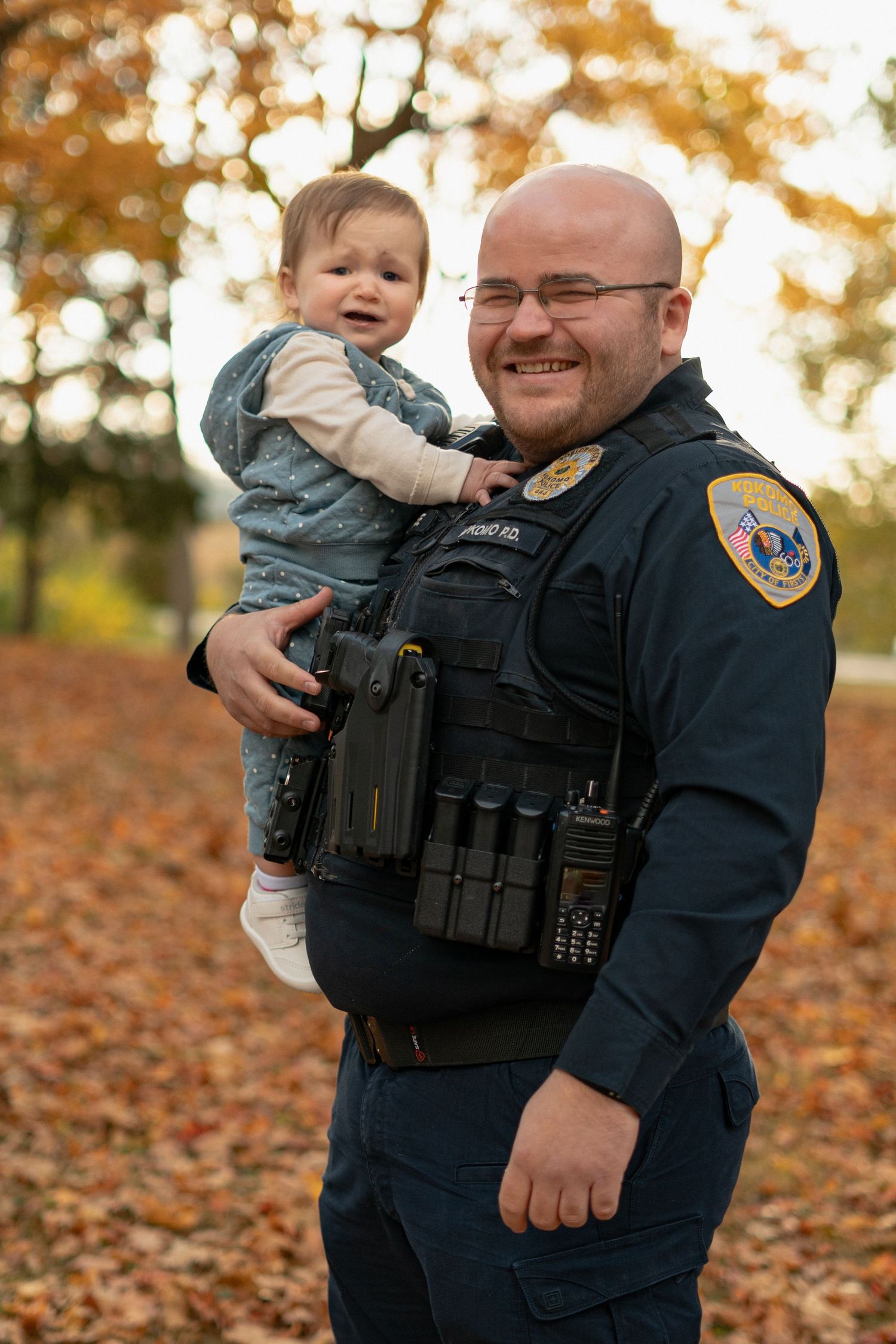 Police officer in uniform holding small child surrounded by autumn foliage.