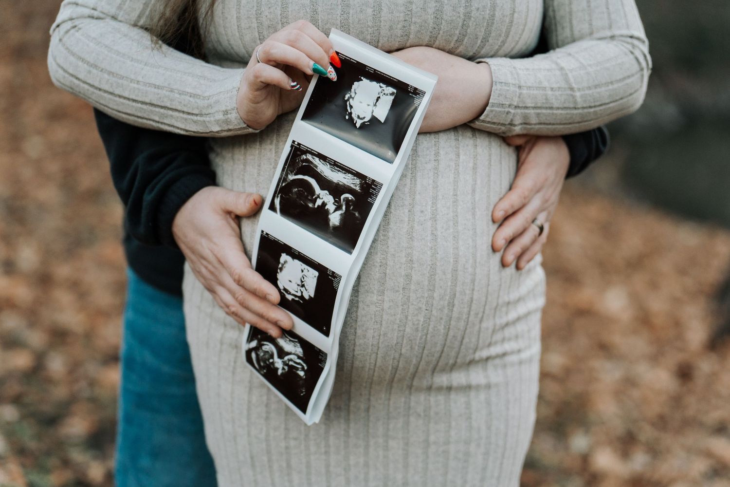 Expectant couple holding ultrasound photos while embracing outdoors during autumn.