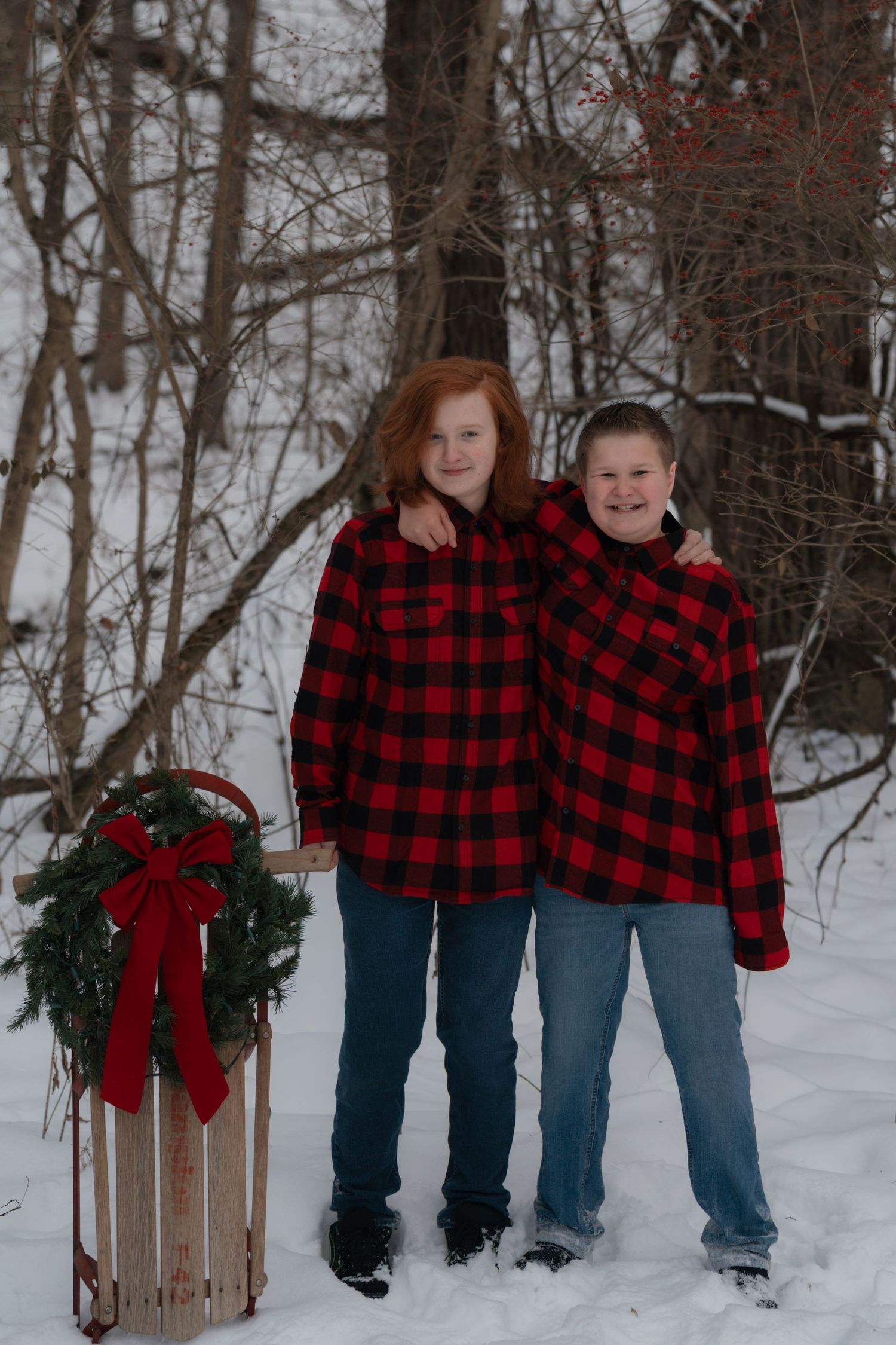 Two people in matching buffalo plaid jackets pose in a snowy winter scene with a decorated wooden sled.