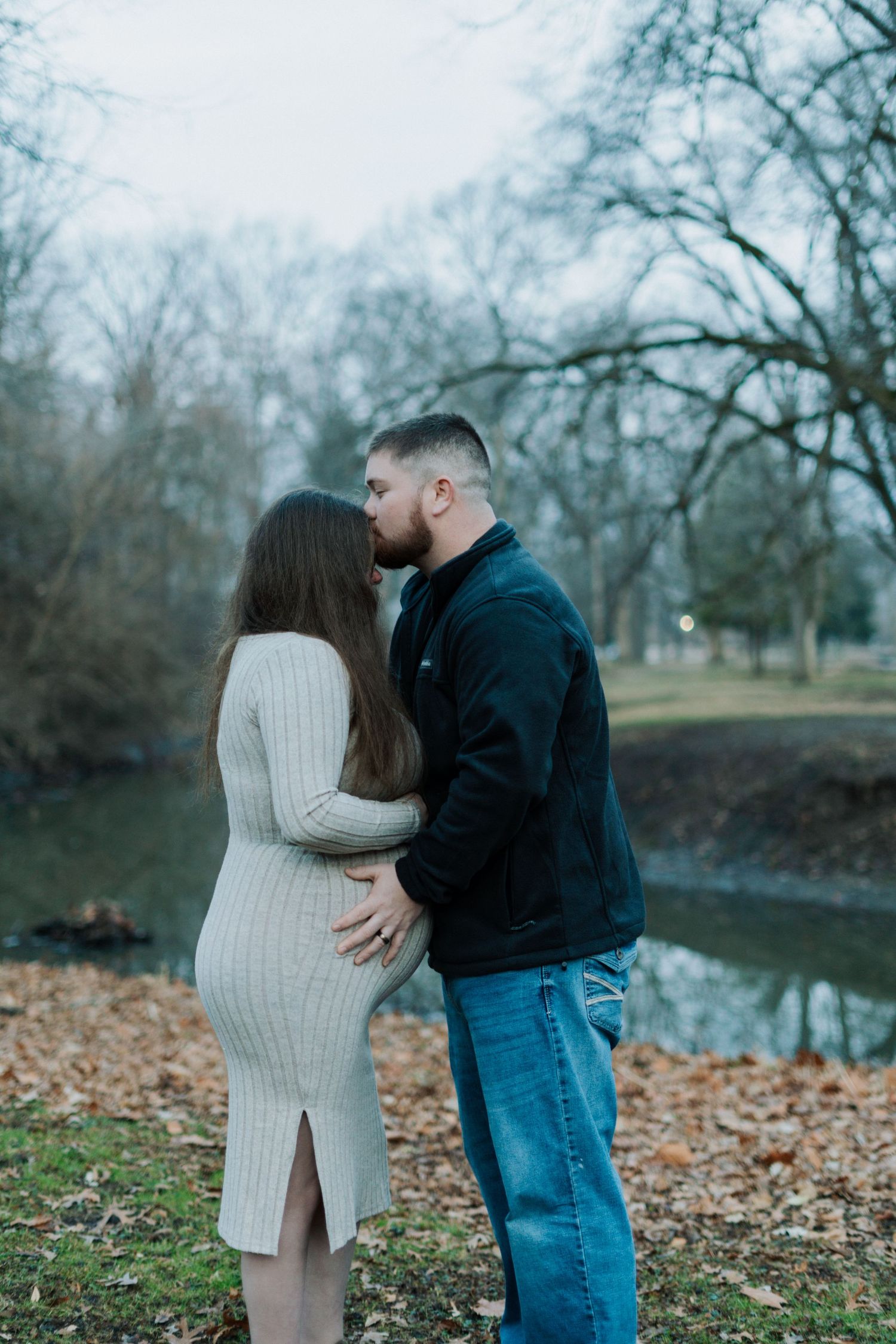 A couple embraces near a serene pond with fallen leaves scattered on the ground.