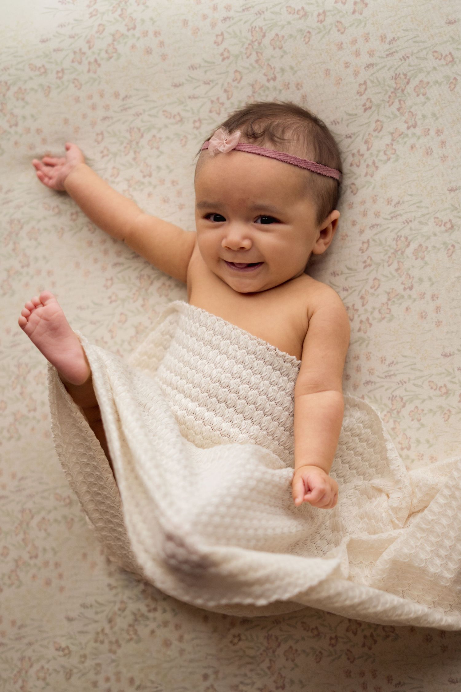 Sweet baby wearing pink headband lies on cream textured blanket while smiling and reaching out.