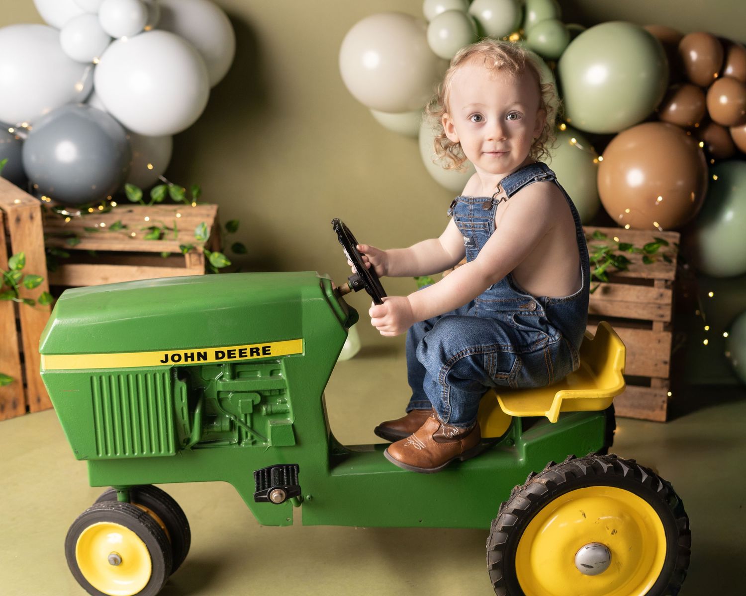 A child sits on a green John Deere toy tractor with festive balloons decorating the background.