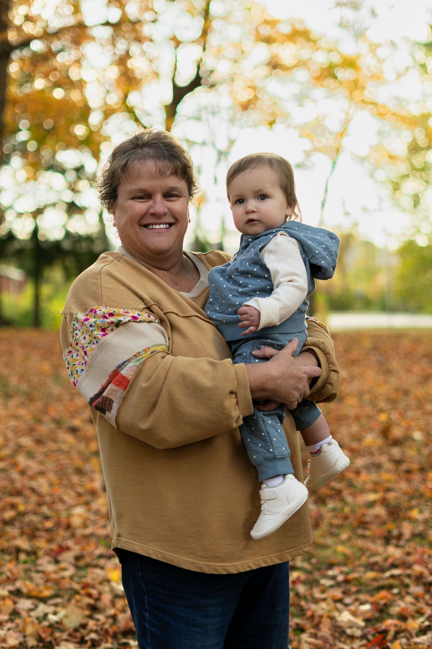 Person in tan sweater holding small child against backdrop of fall leaves.
