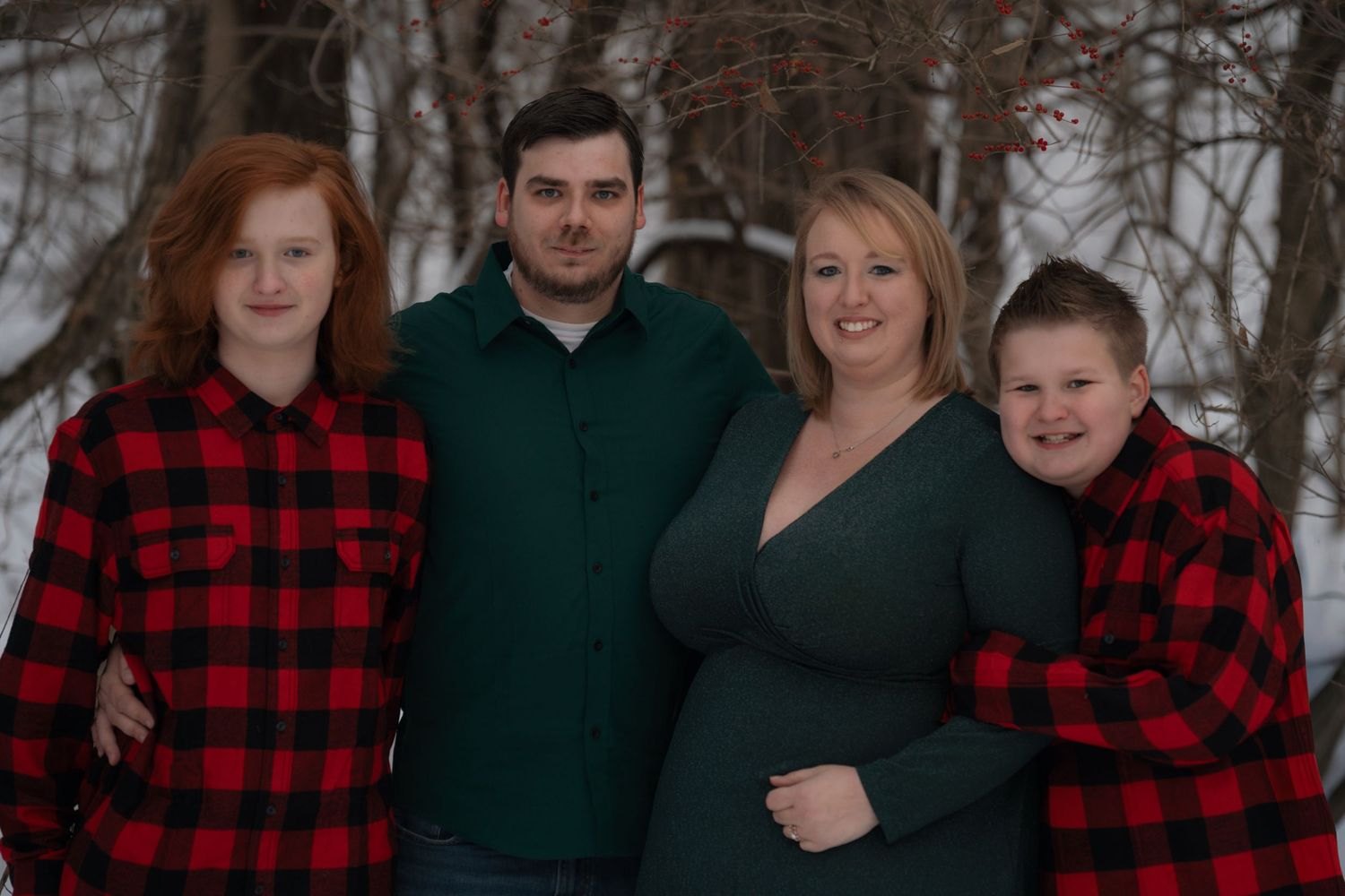A family wearing coordinated red buffalo plaid and dark green outfits poses together for outdoor winter portraits.