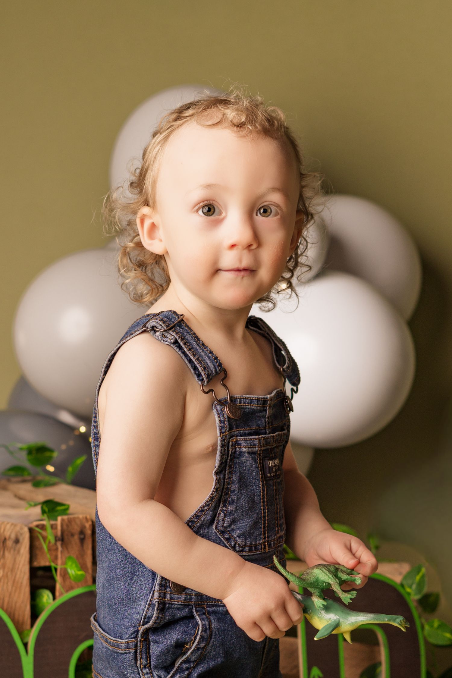 A toddler in denim overalls plays with props against a neutral background with white balloons.