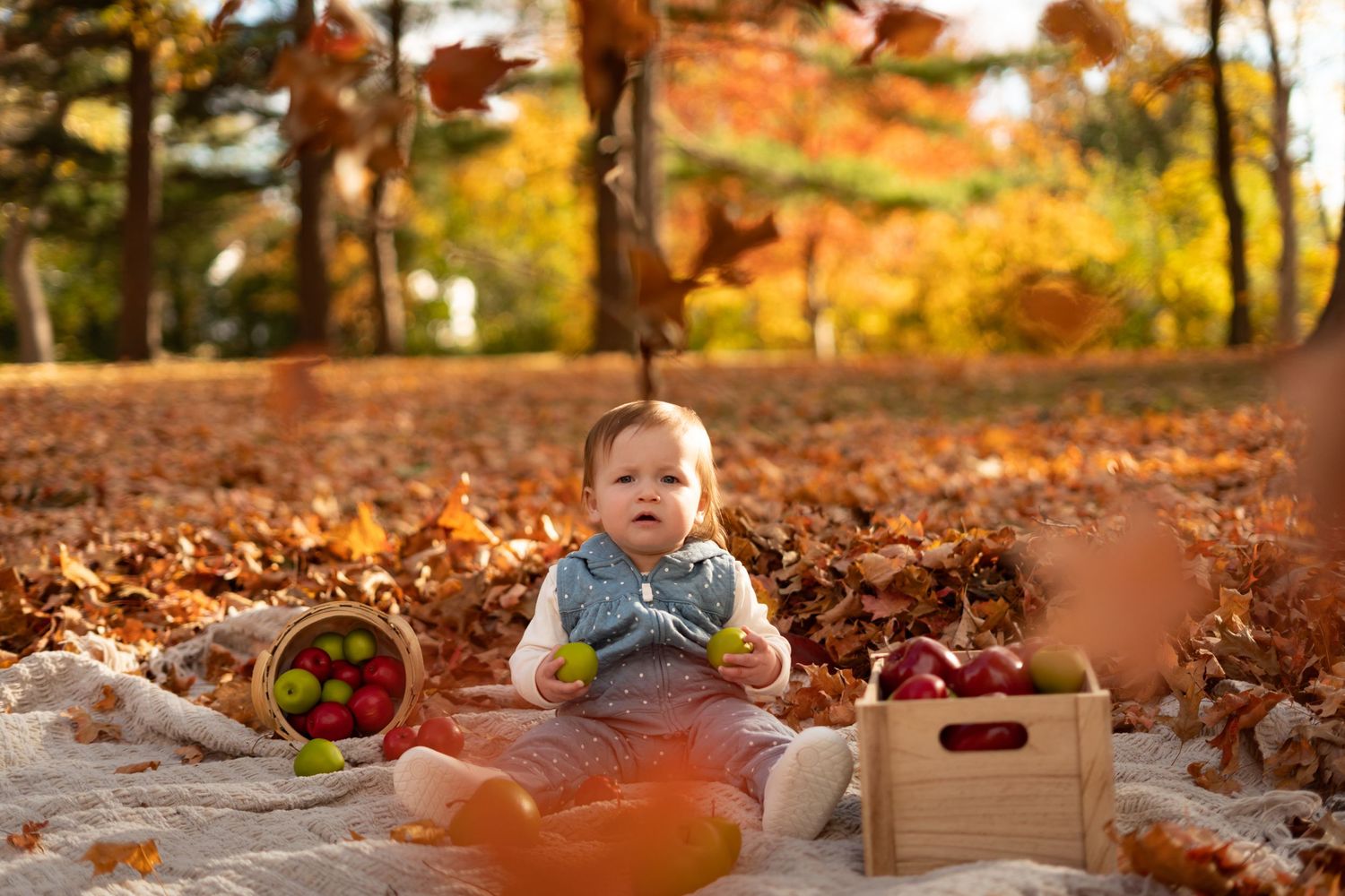 Baby sitting on white blanket with wooden crates of apples during autumn photo session.