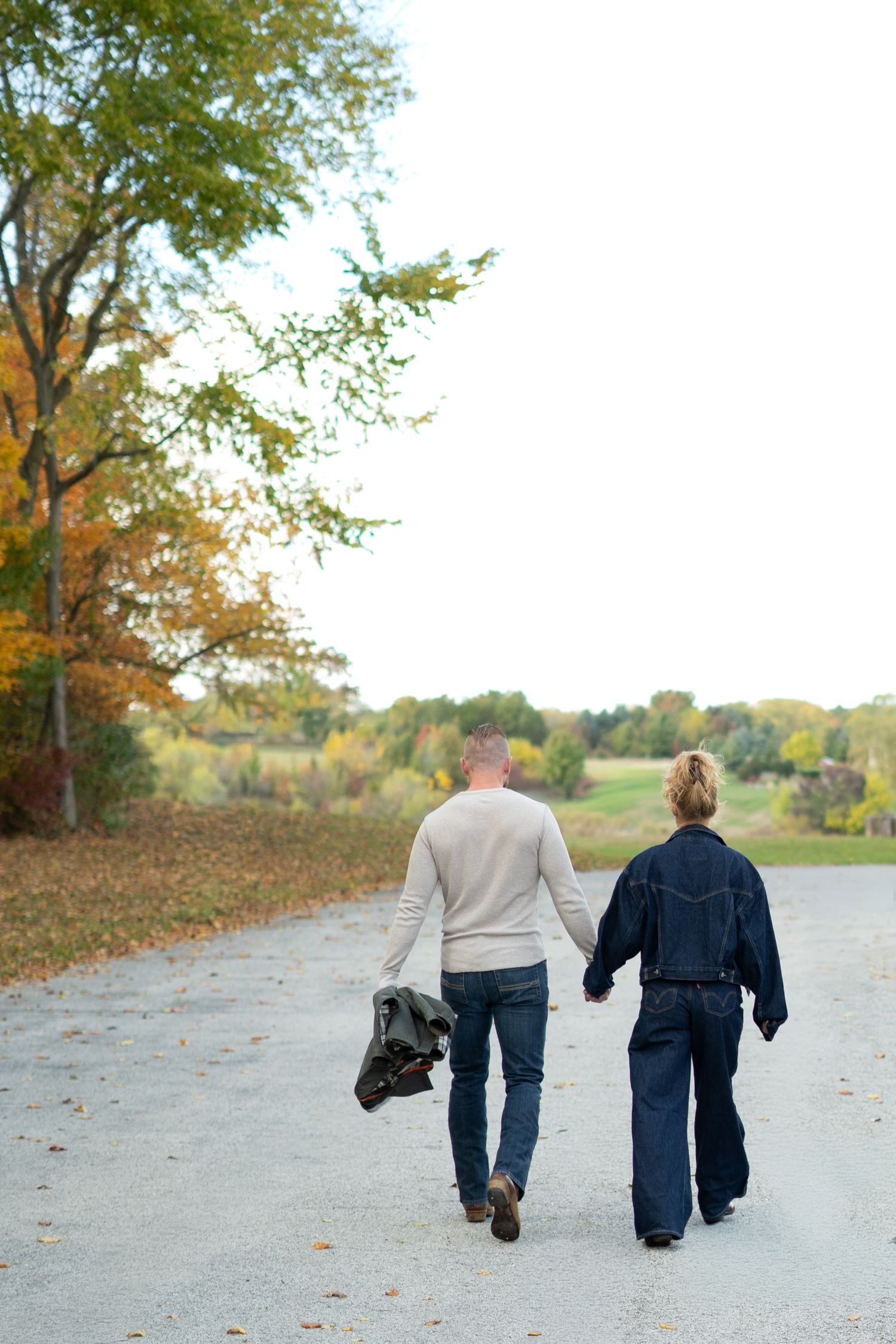 A couple walks away together on a path surrounded by autumn trees and fallen leaves.