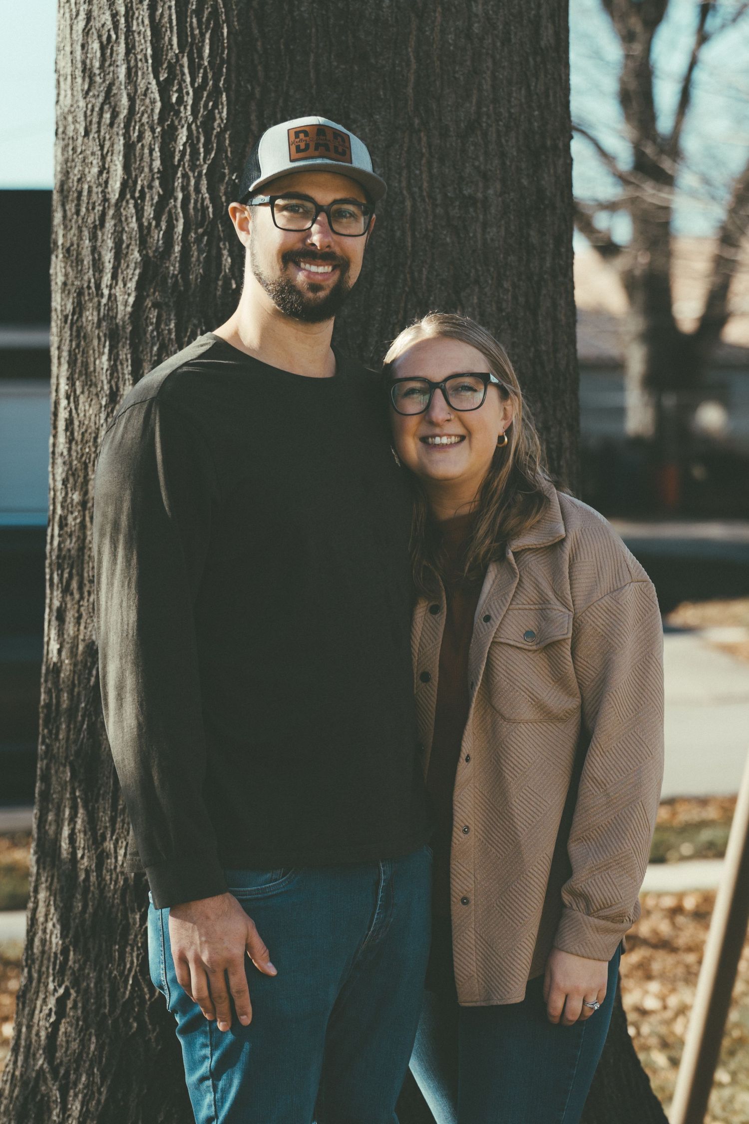 Couple posing together near a large tree trunk in autumn wearing casual outfits.