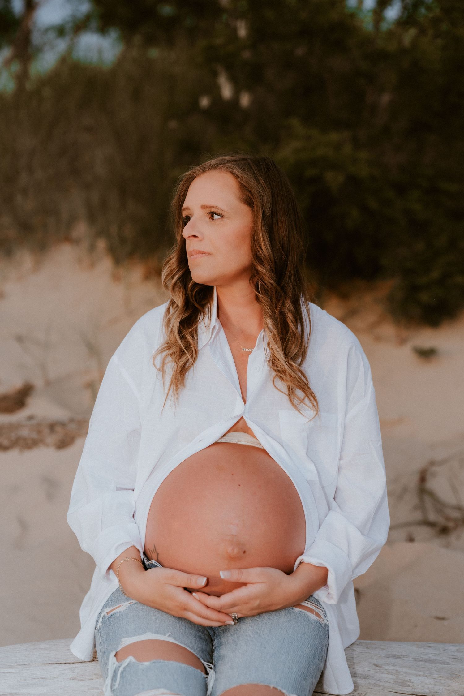 Pregnant woman in white shirt and ripped jeans cradling baby bump while sitting on beach for maternity photography session North Central IN