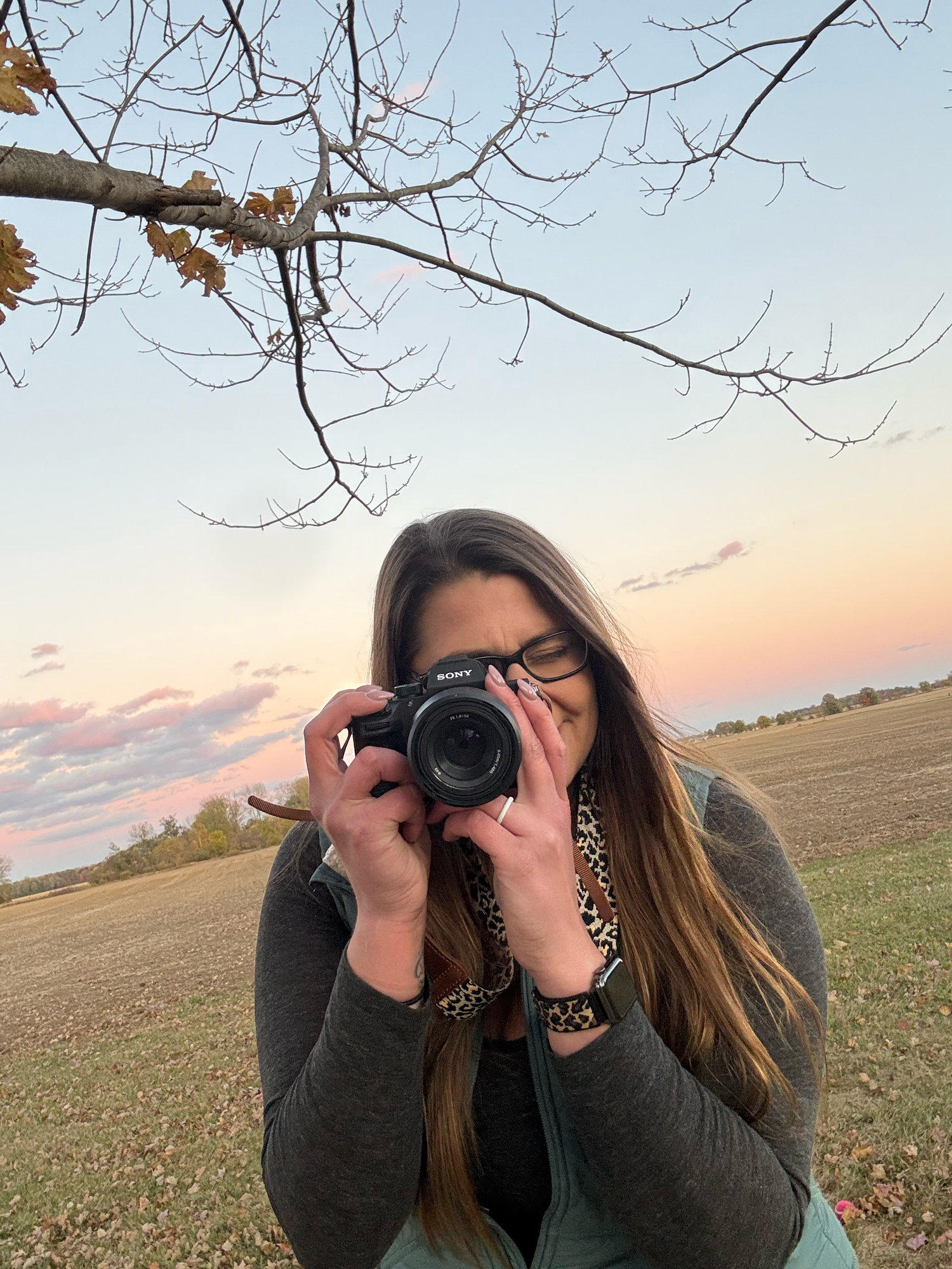 Photographer with long hair taking pictures in a rural field during sunset with bare tree branches overhead.
