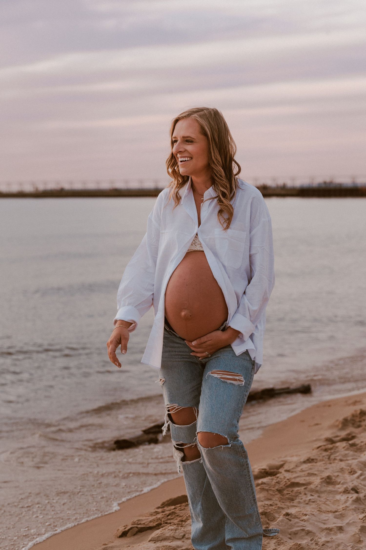 Smiling pregnant woman in white shirt and ripped jeans cradling baby bump on sandy beach during maternity photo session in North Central IN