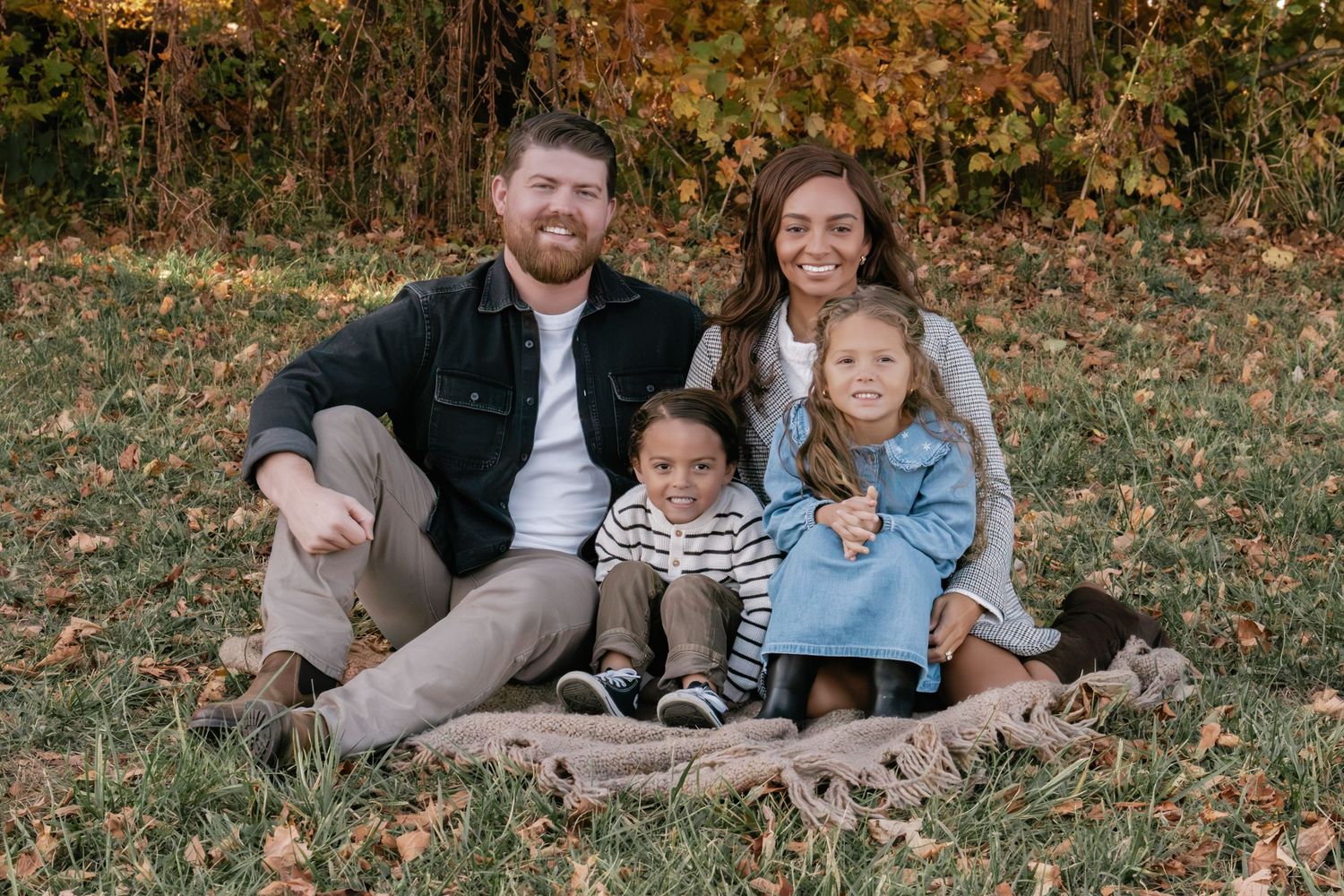 Family relaxes together on fallen leaves during autumn photoshoot in wooded setting.