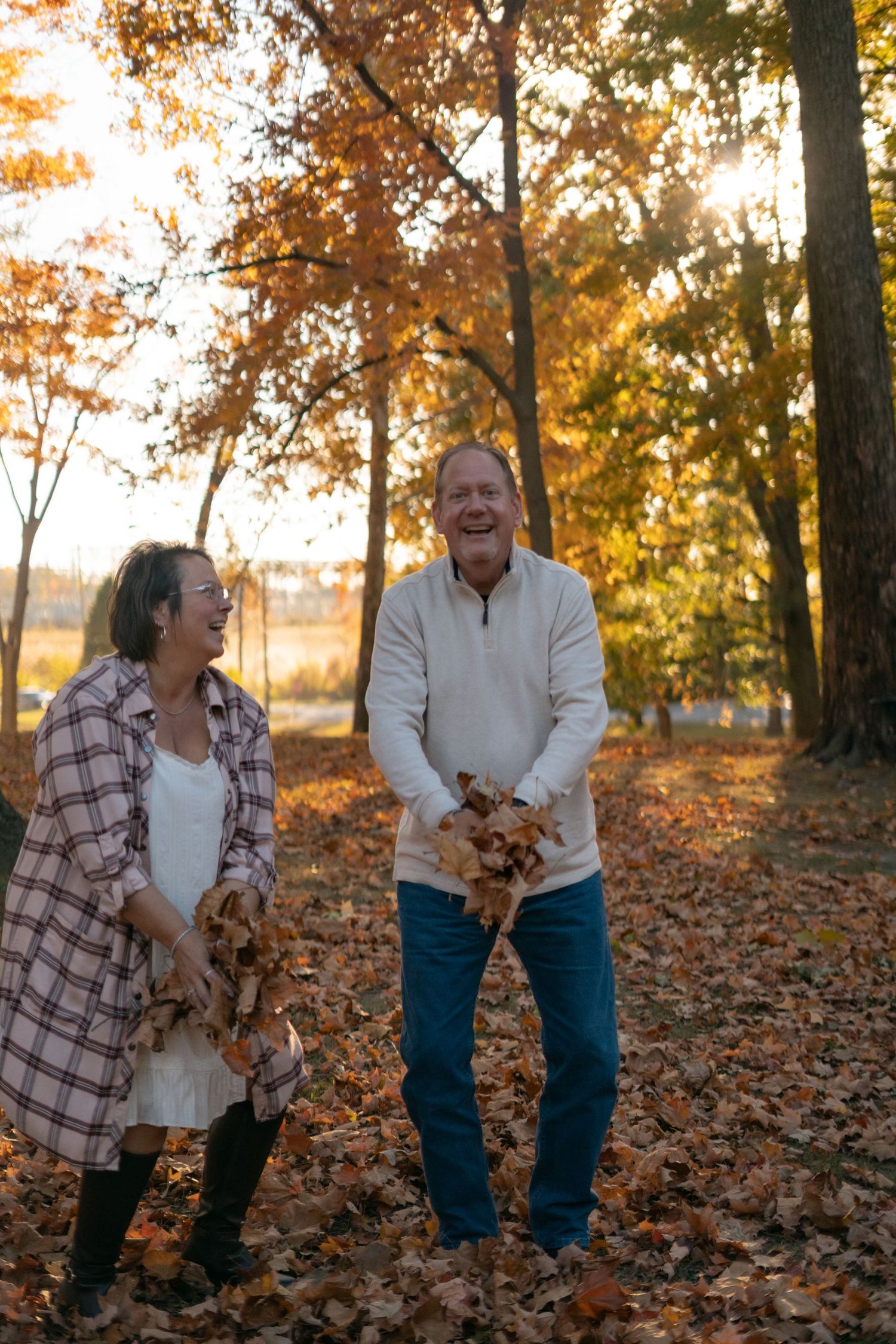 Two people laugh while playing with fallen leaves in an autumn park.