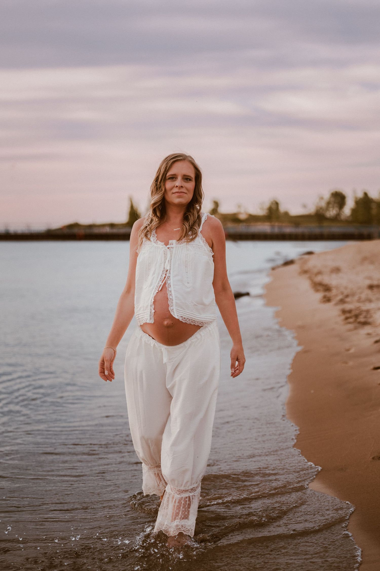 Pregnant woman in white outfit walking along sandy beach shoreline at sunset during maternity photo session in North Central IN