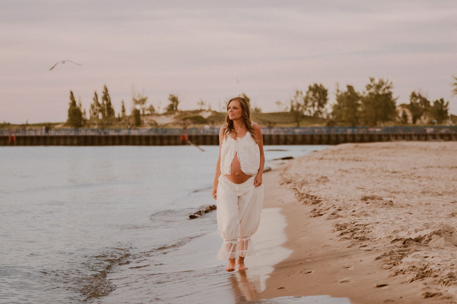 Pregnant woman in white outfit walking along sandy beach at golden hour sunset near pier during maternity photo session in North Central IN