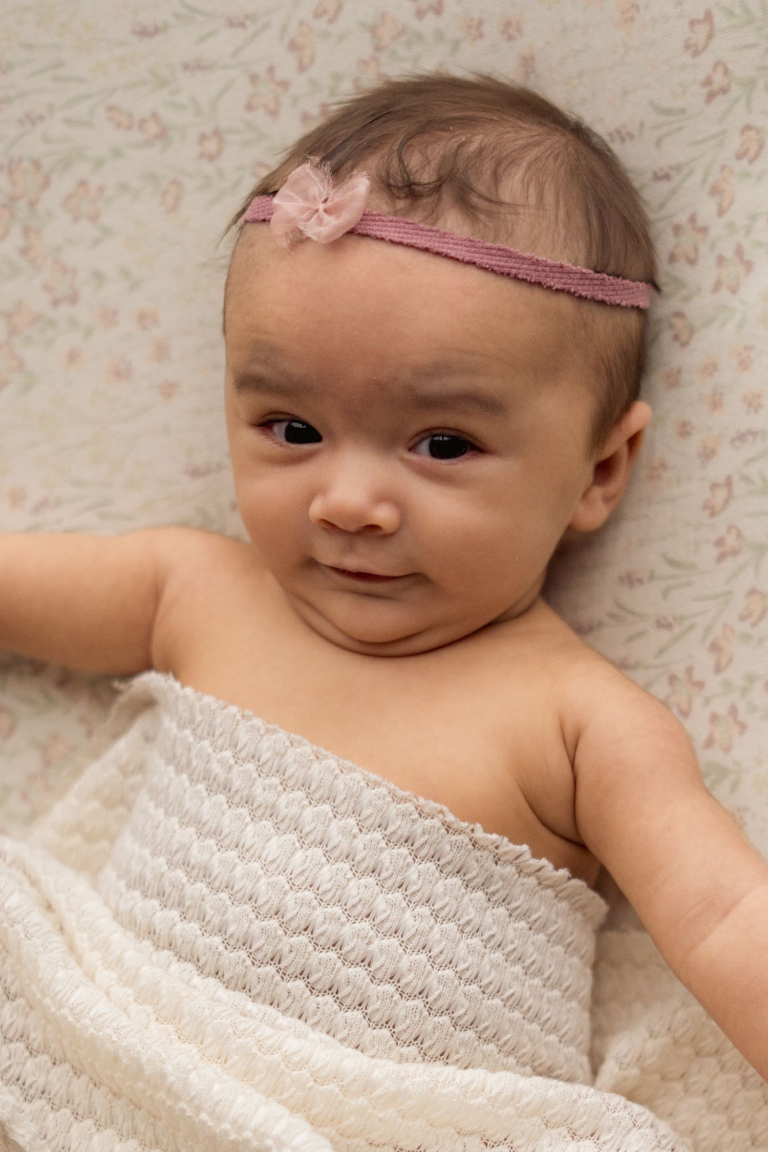 Baby wearing pink headband lies on floral background wrapped in white textured blanket.