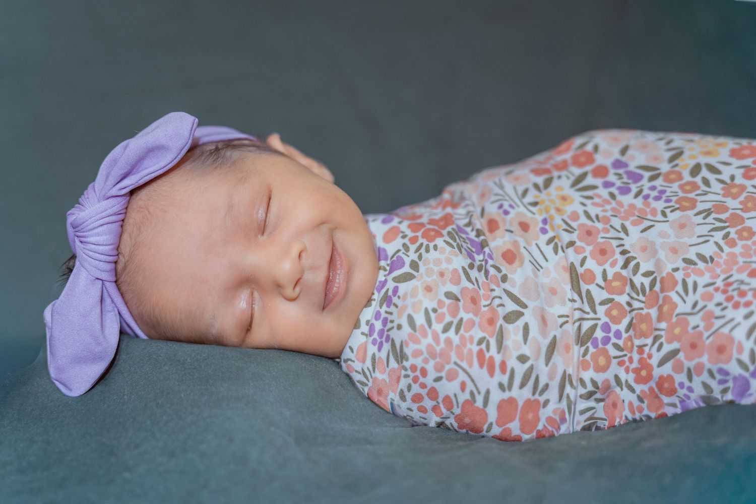 Newborn baby sleeping peacefully in floral swaddle with purple bow headband.
