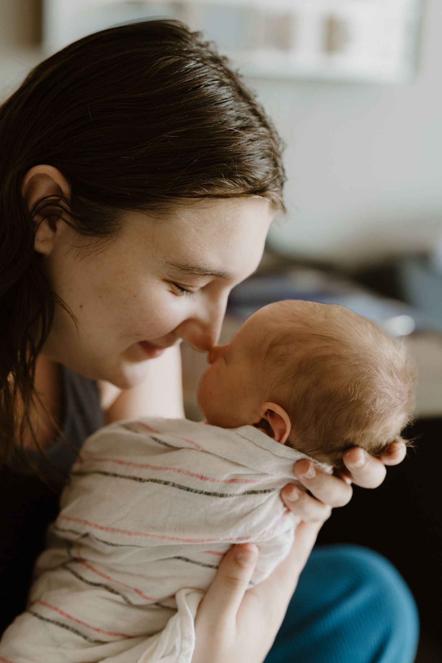 A young mother tenderly touches noses with her newborn baby wrapped in a striped muslin swaddle blanket during in hospital Fresh 48 Indiana.