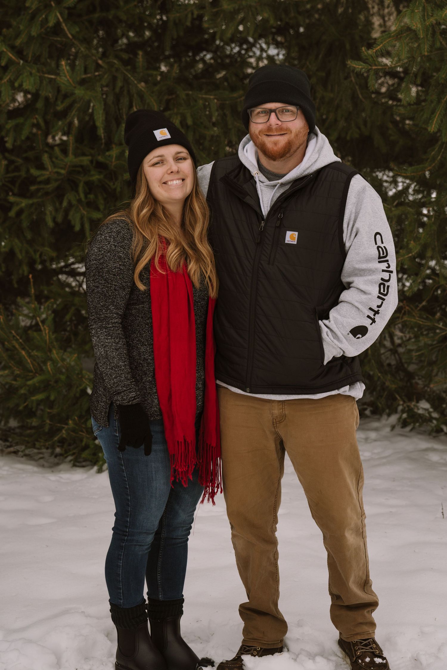 Couple standing together in snow wearing winter coats and a bright red scarf.