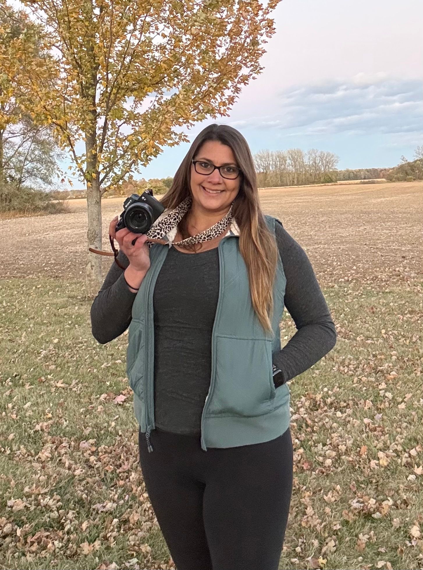 A photographer holds a camera while standing in a fall field with autumn trees in the background.