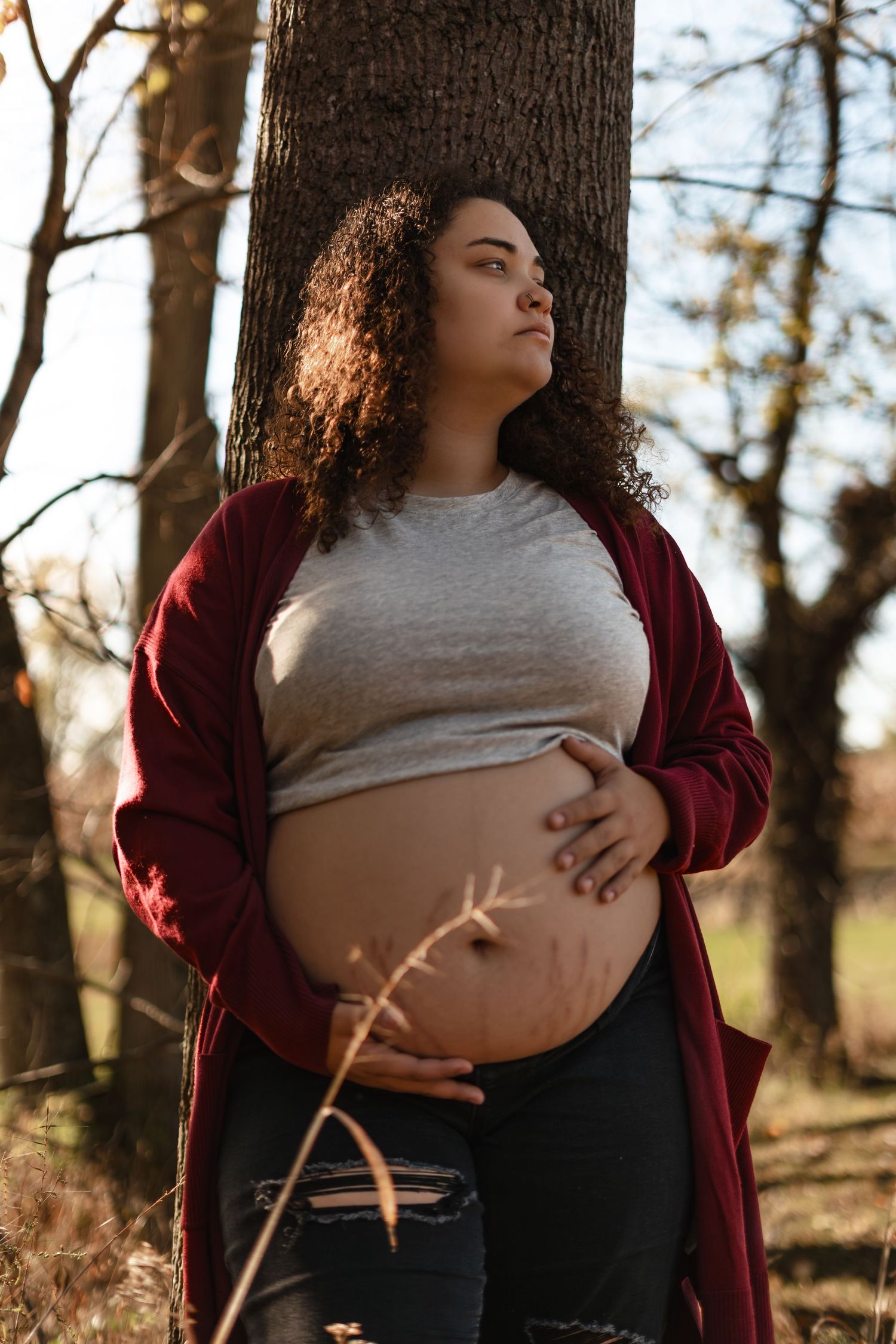 Maternity portrait against a tree trunk with burgundy cardigan and natural lighting.