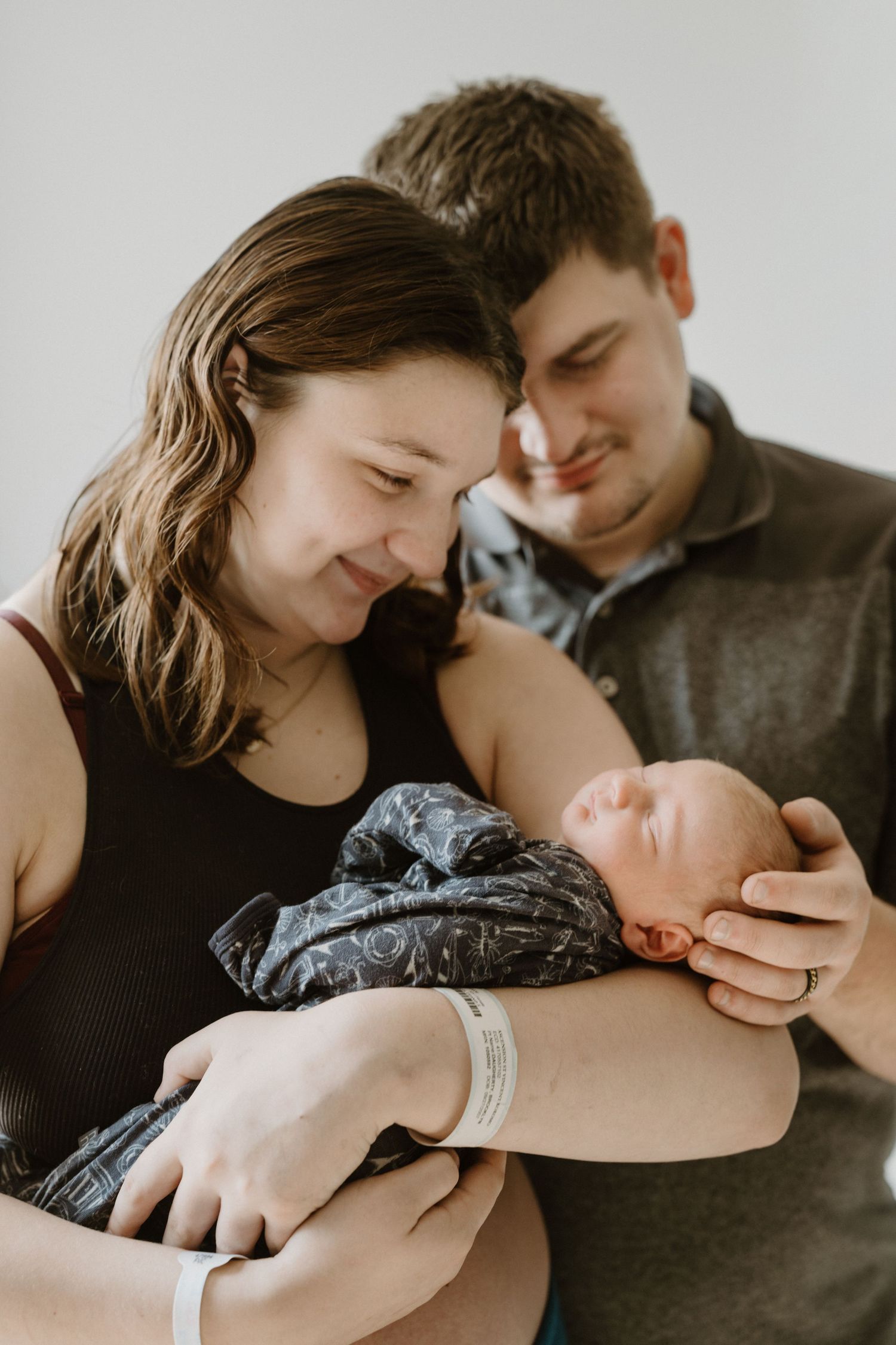 In-home newborn photography session with new parents cradling their sleeping baby wrapped in a dark swaddle in North Central Indiana.