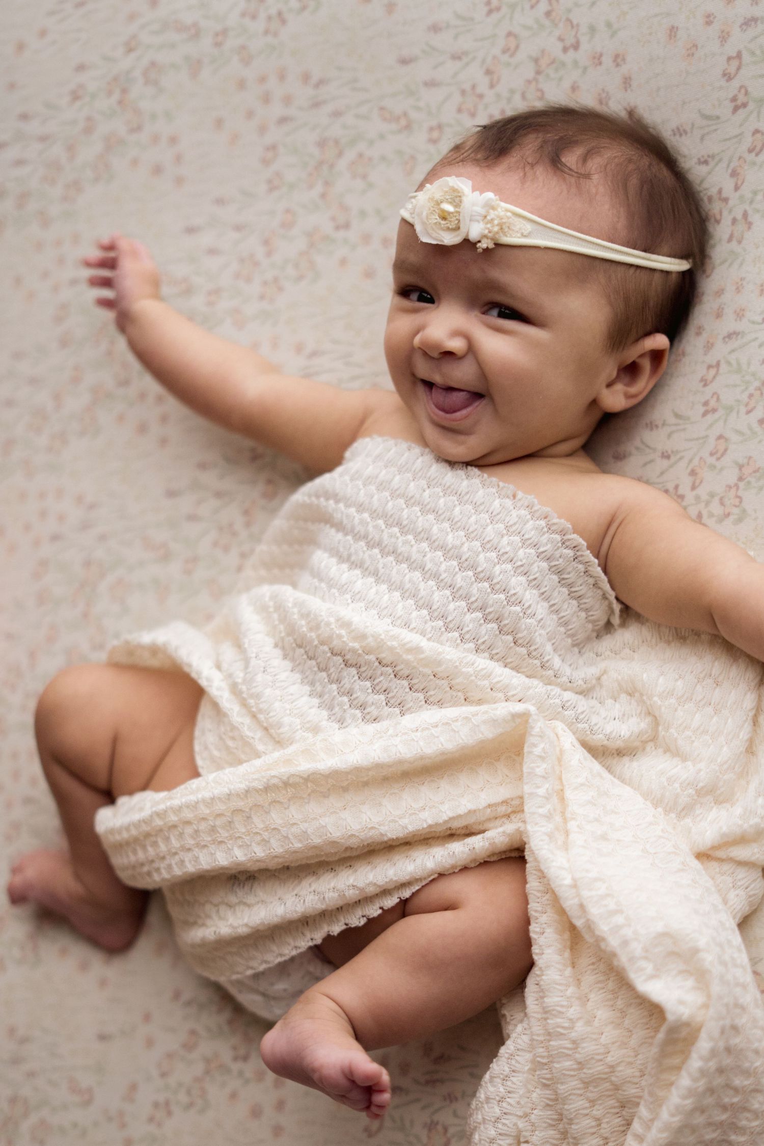 Happy baby in white knit dress smiles while lying on floral patterned fabric.
