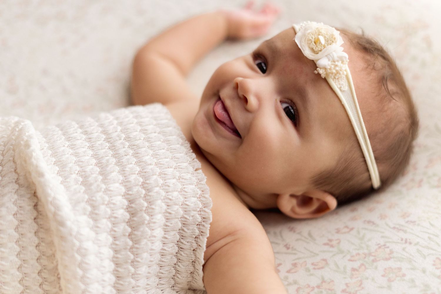 Baby wearing delicate lace headband smiles while lying on soft textured blanket.