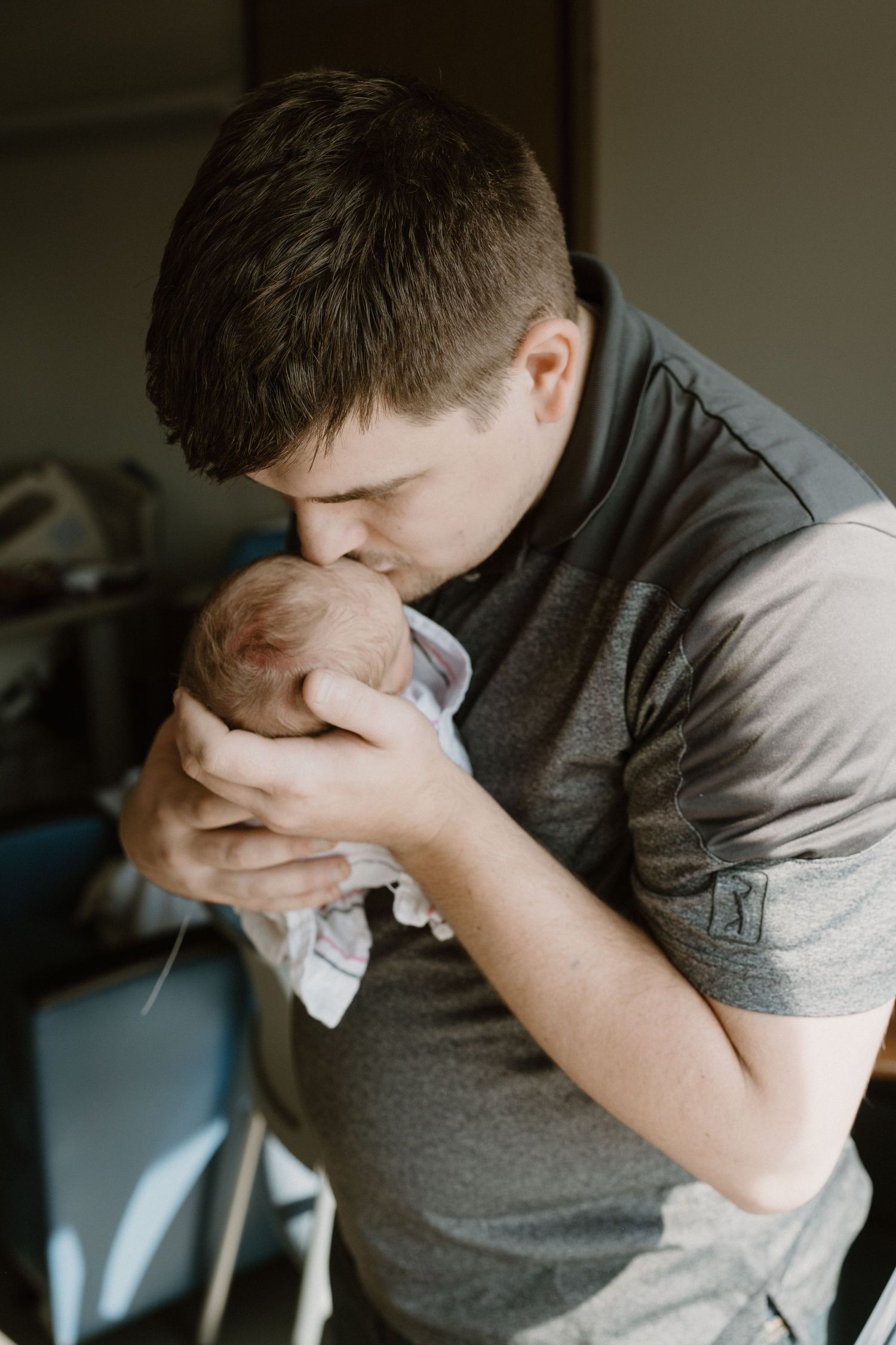 A young father tenderly kisses his newborn baby while cradling the infant gently in both hands during in-hospital newborn session Indiana