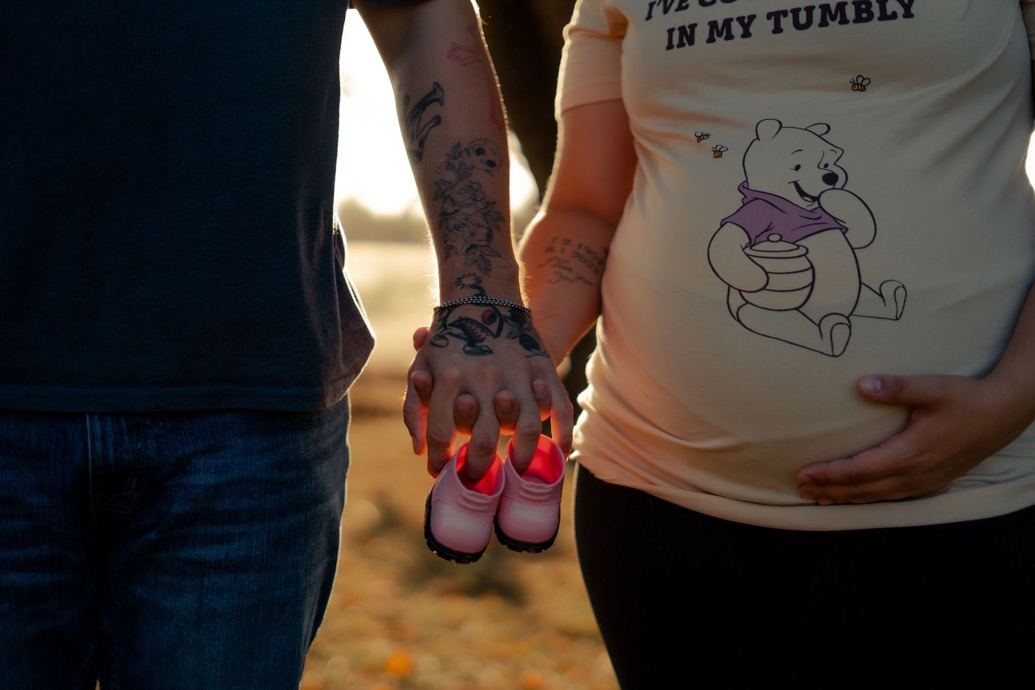 A couple holding tiny pink baby shoes at sunset, with a Winnie the Pooh maternity shirt visible.