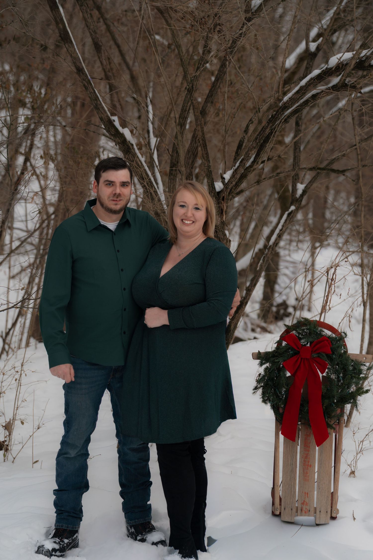Couple in matching outfits posing with holiday wreaths in a snowy winter setting.