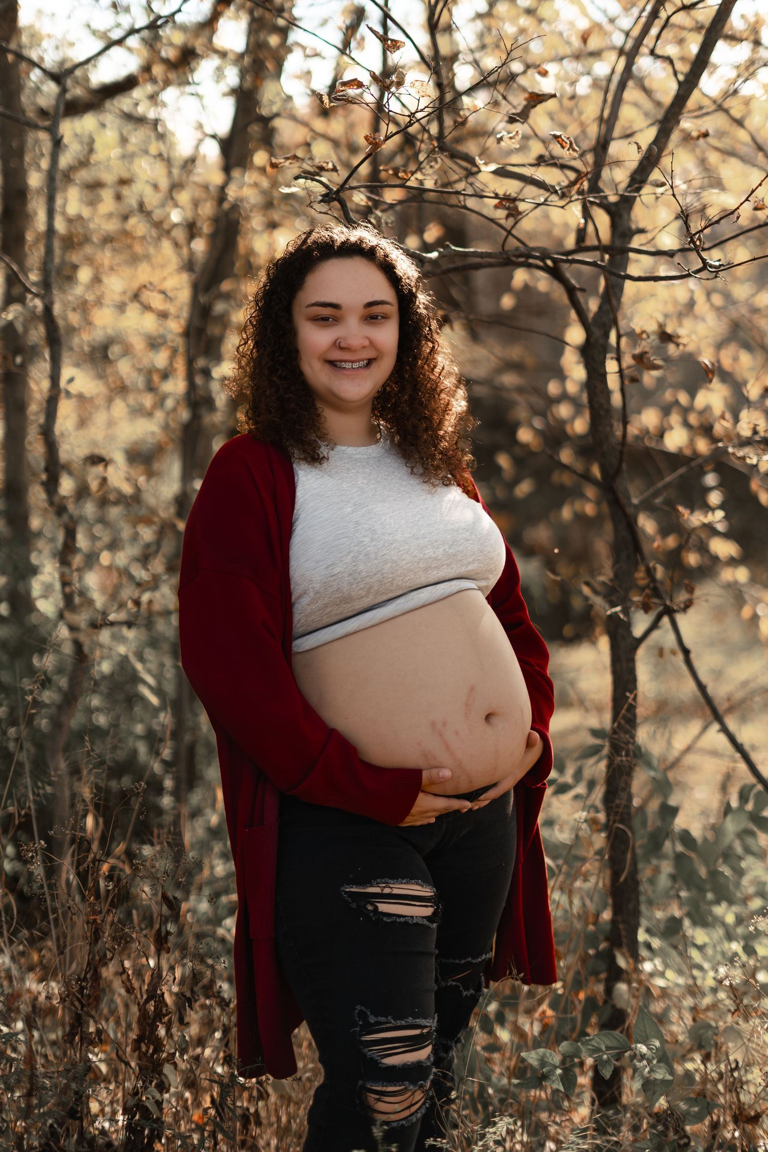 A maternity photo in nature featuring burgundy cardigan and ripped jeans in autumn setting.