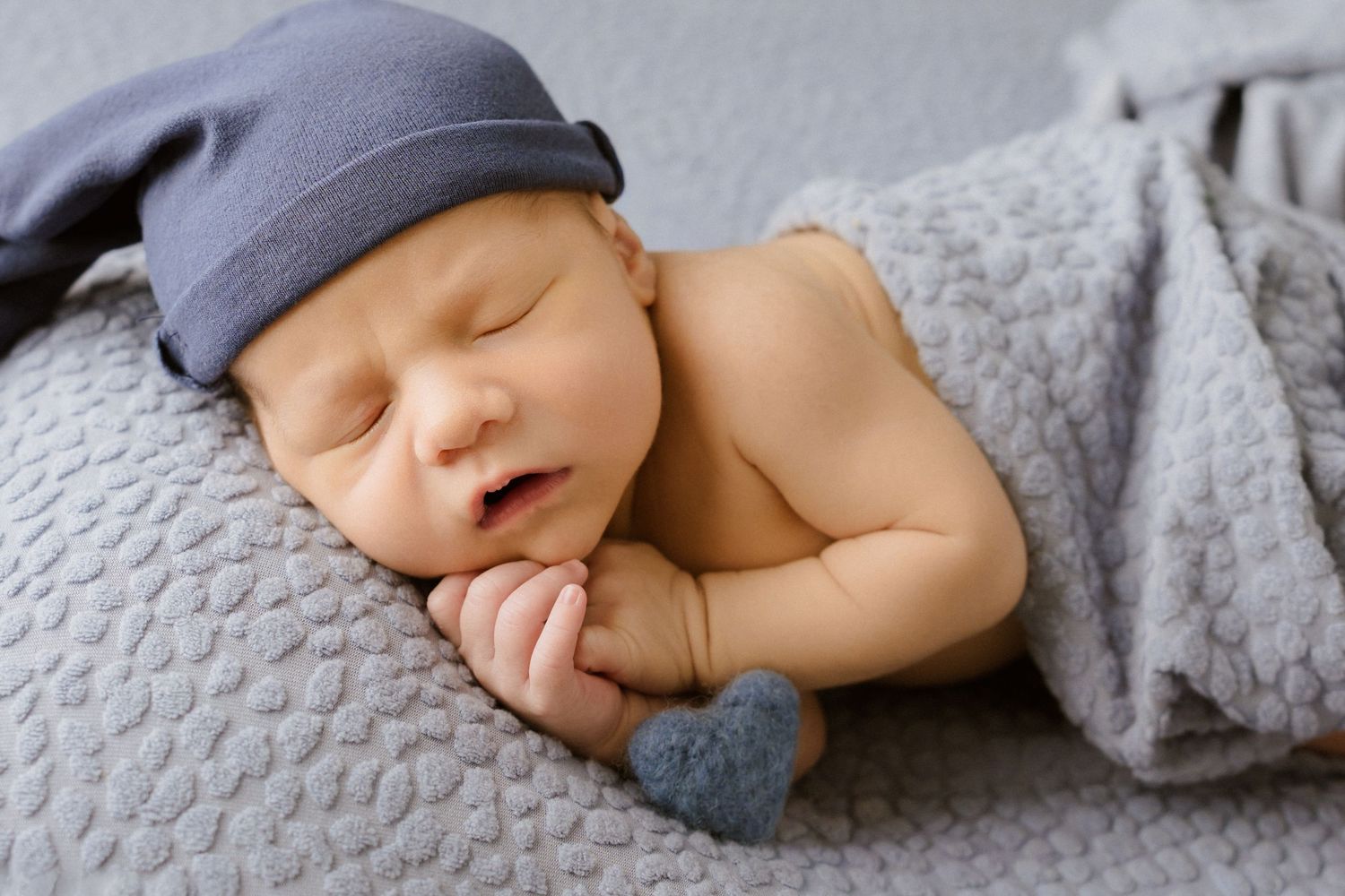 Peaceful newborn baby sleeping on gray blanket wearing blue hat, curled up in cozy pose during in-home newborn session North Central IN
