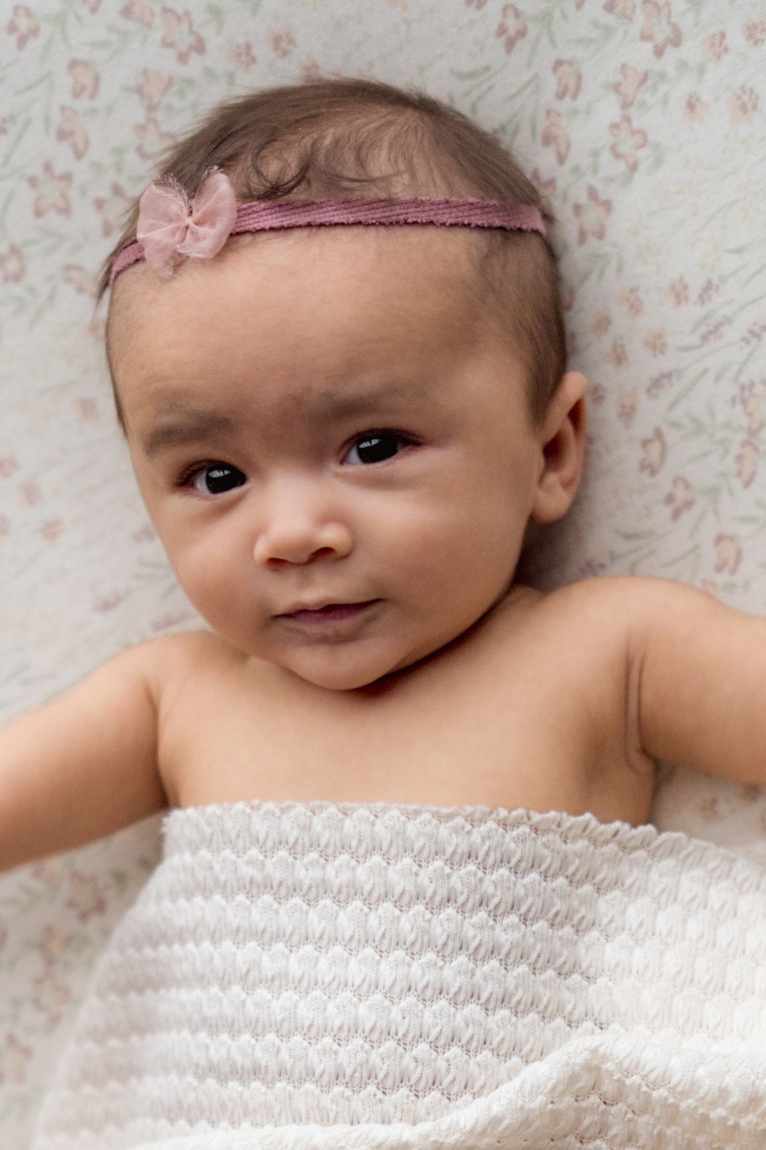 Sweet newborn baby wearing a pink headband lies on floral bedding while smiling at the camera.