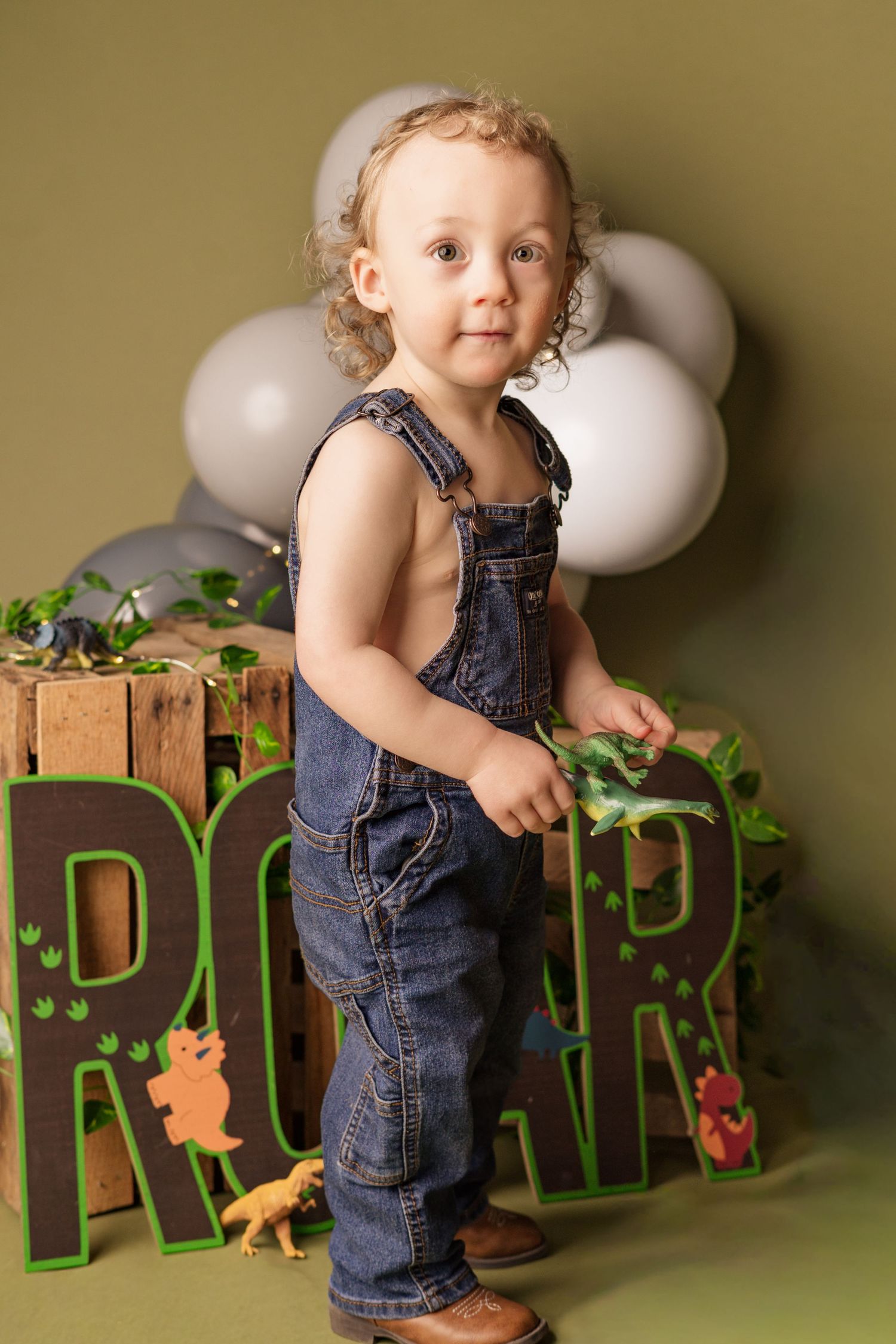 Toddler wearing denim overalls stands in front of white balloons and decorative green letters with dinosaur accents.