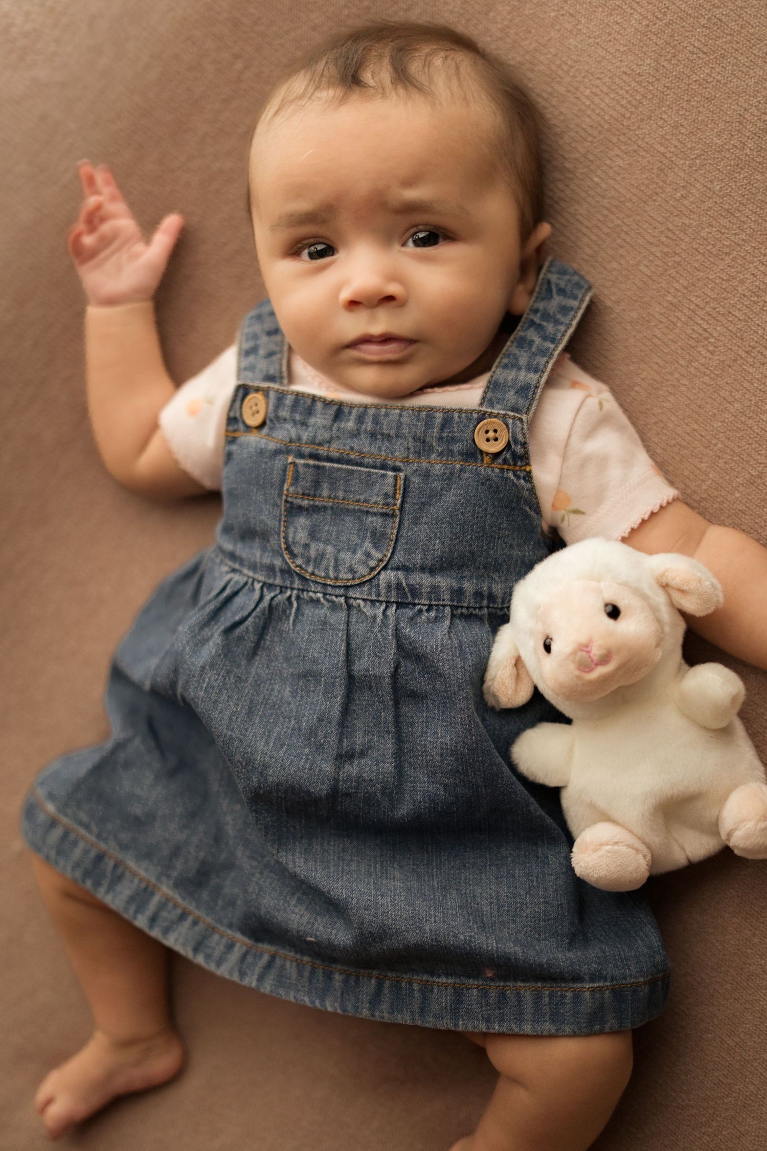 Baby in denim dress lying on brown backdrop with white plush bunny toy.