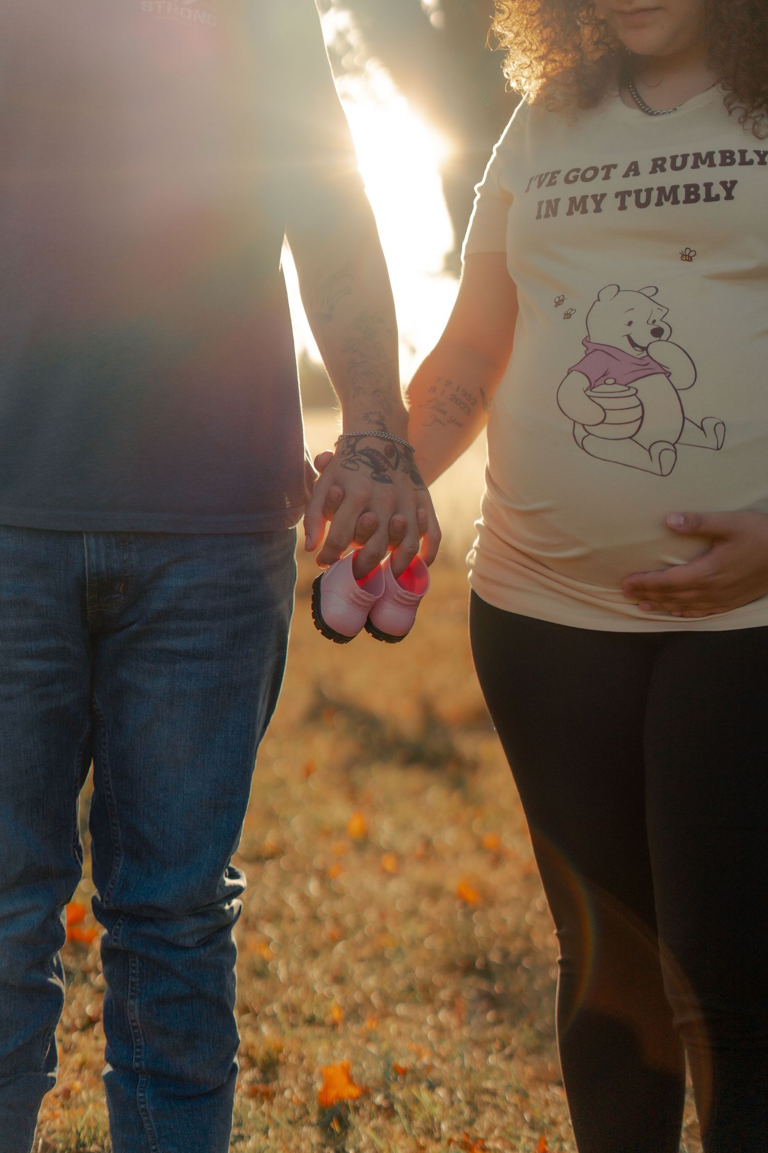 Couple holding pink baby shoes against sunset with Winnie the Pooh maternity shirt visible in warm lighting.