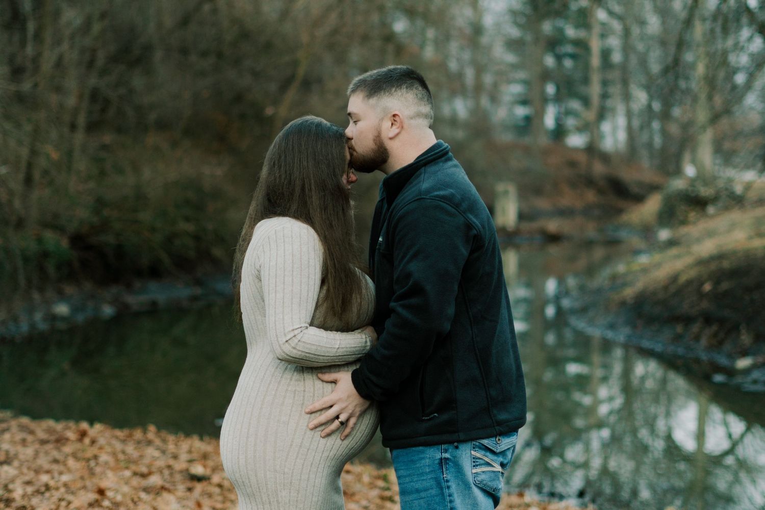 A couple embraces near a tranquil pond with fallen leaves scattered on the ground.