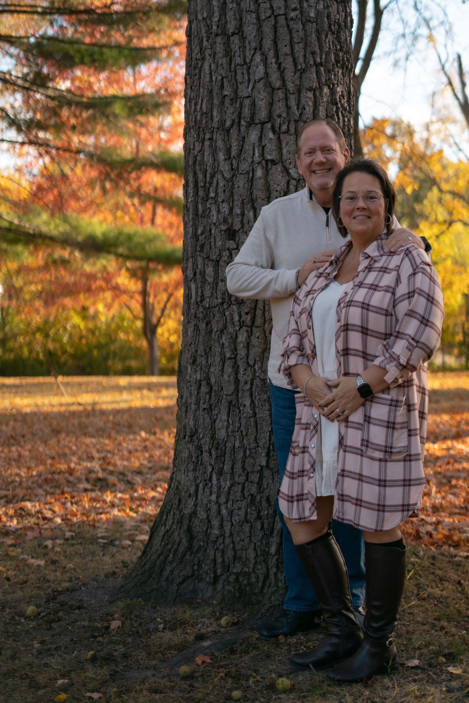 Two people pose together against a large tree trunk in autumn scenery.